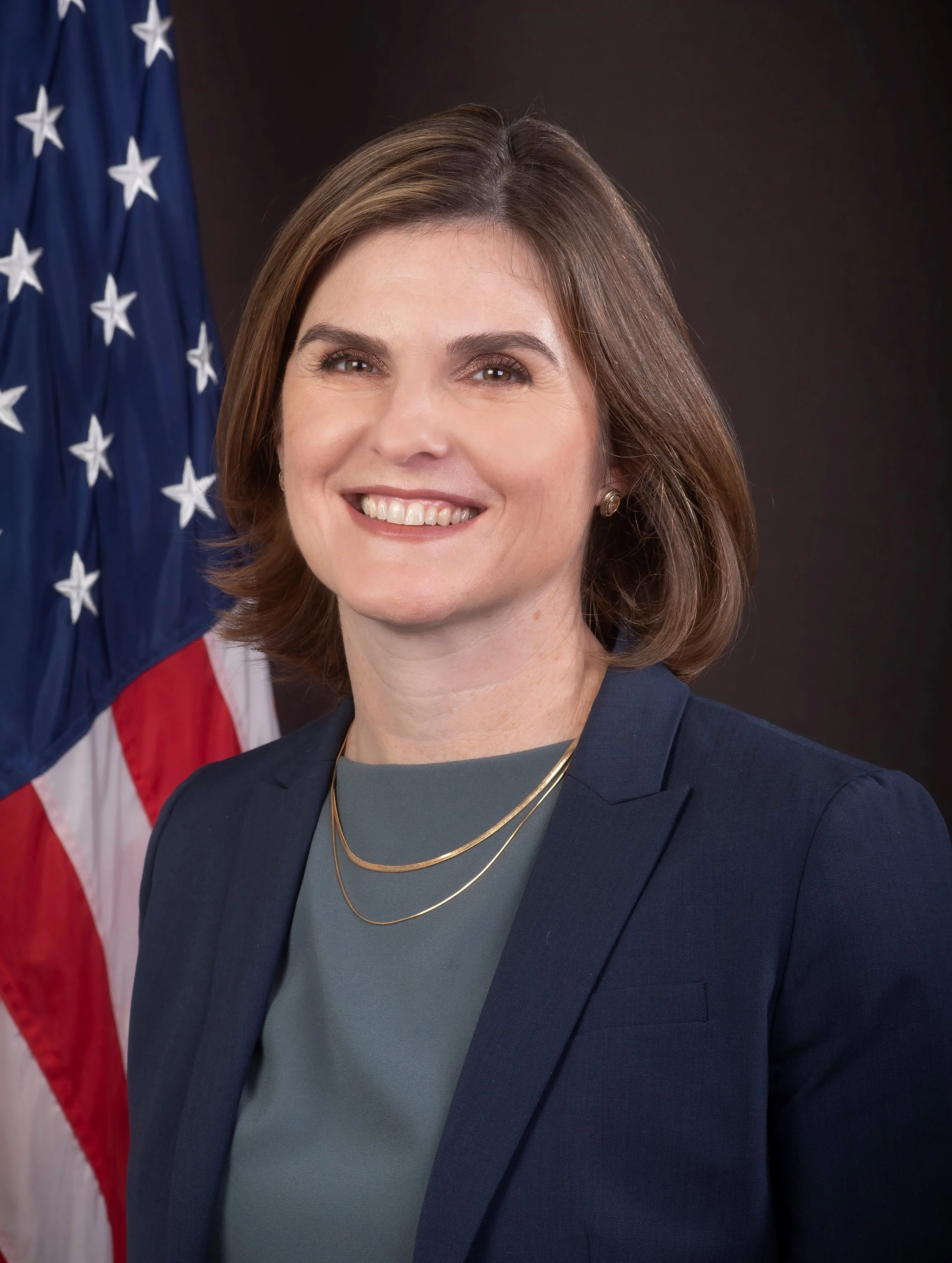 A woman with shoulder-length brown hair smiling in front of the American flag, wearing a navy blazer, gray top, and layered gold necklaces.