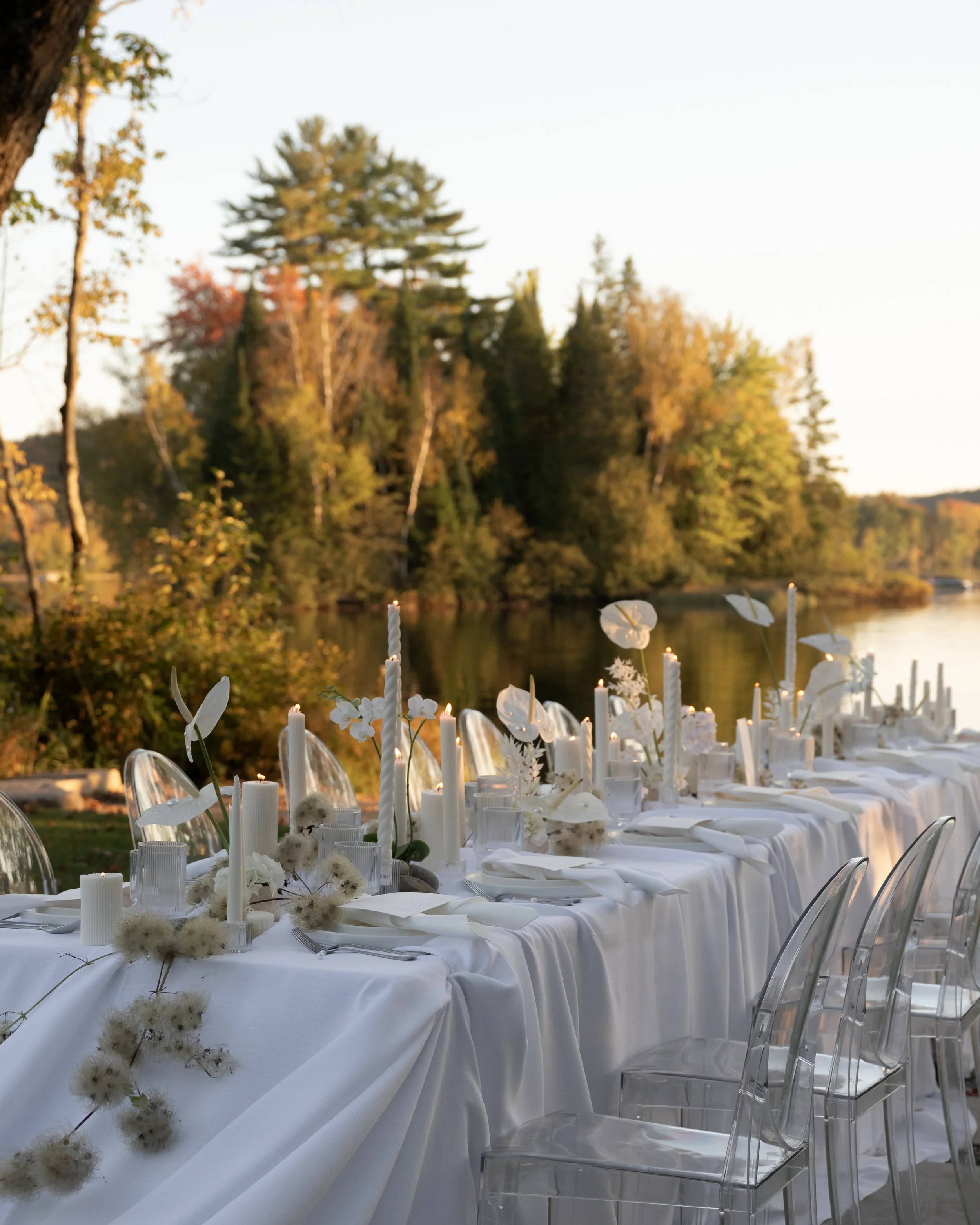 Elegant outdoor dining setup along a river with white tablecloth, white candles, and transparent chairs, surrounded by trees in fall colors.