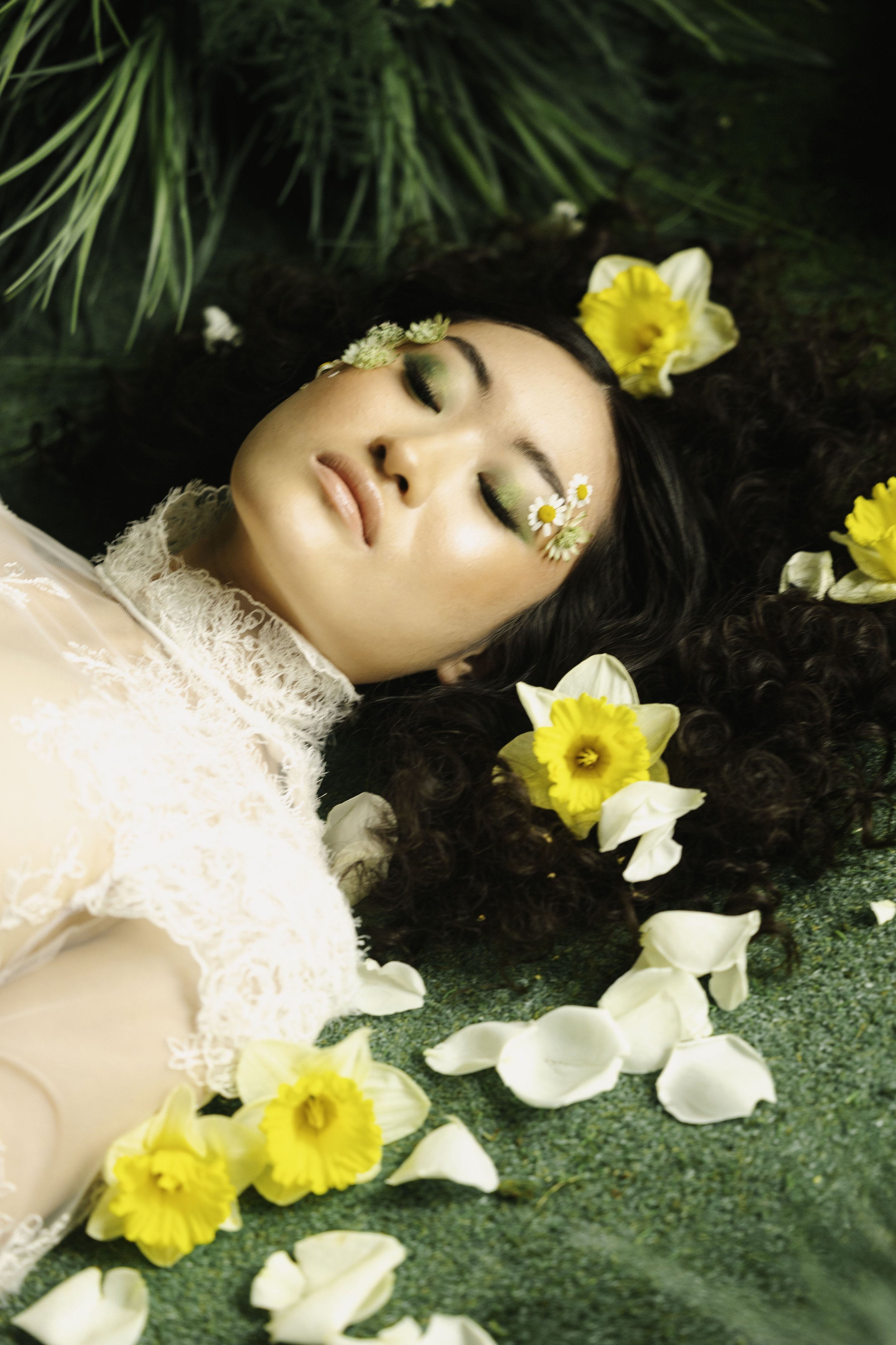 A woman with dark curly hair and floral makeup lies on green grass surrounded by yellow and white flowers, with her eyes closed and wearing a white lace top.