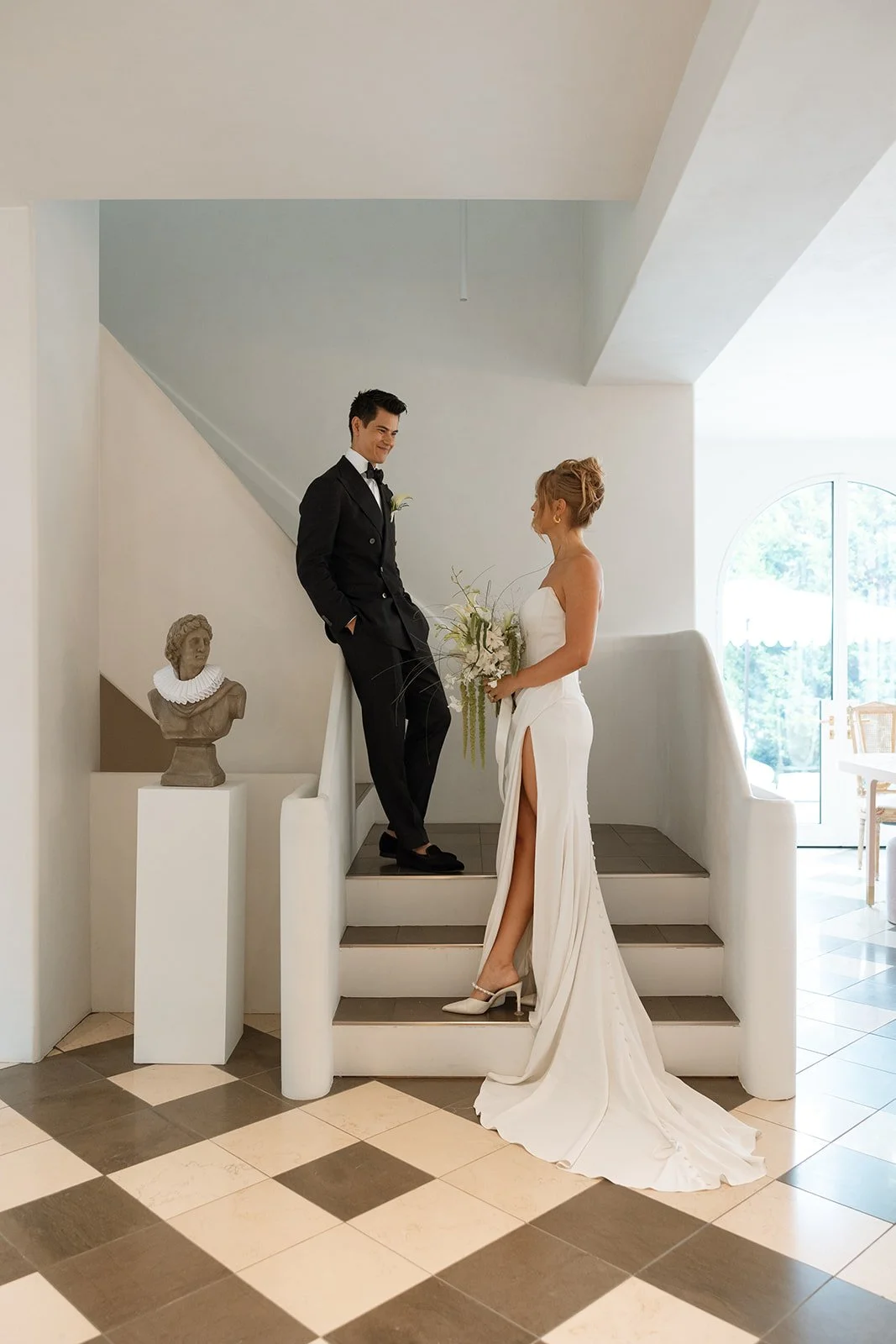 Bride in white gown holding bouquet and groom in black tuxedo on stairs indoors during wedding.