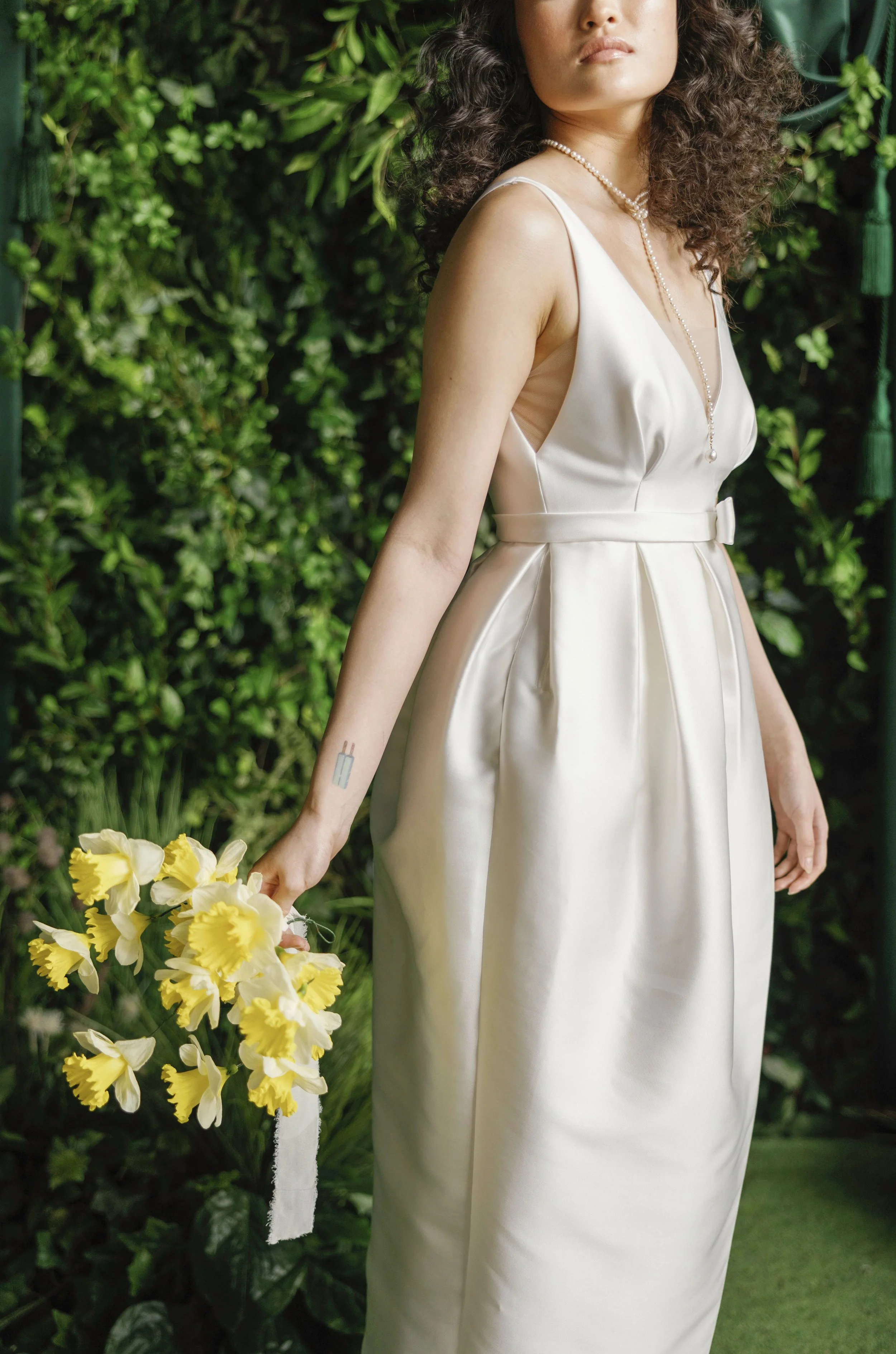 Woman in a satin white dress holding a bunch of yellow daffodils, standing against a lush green leafy background.