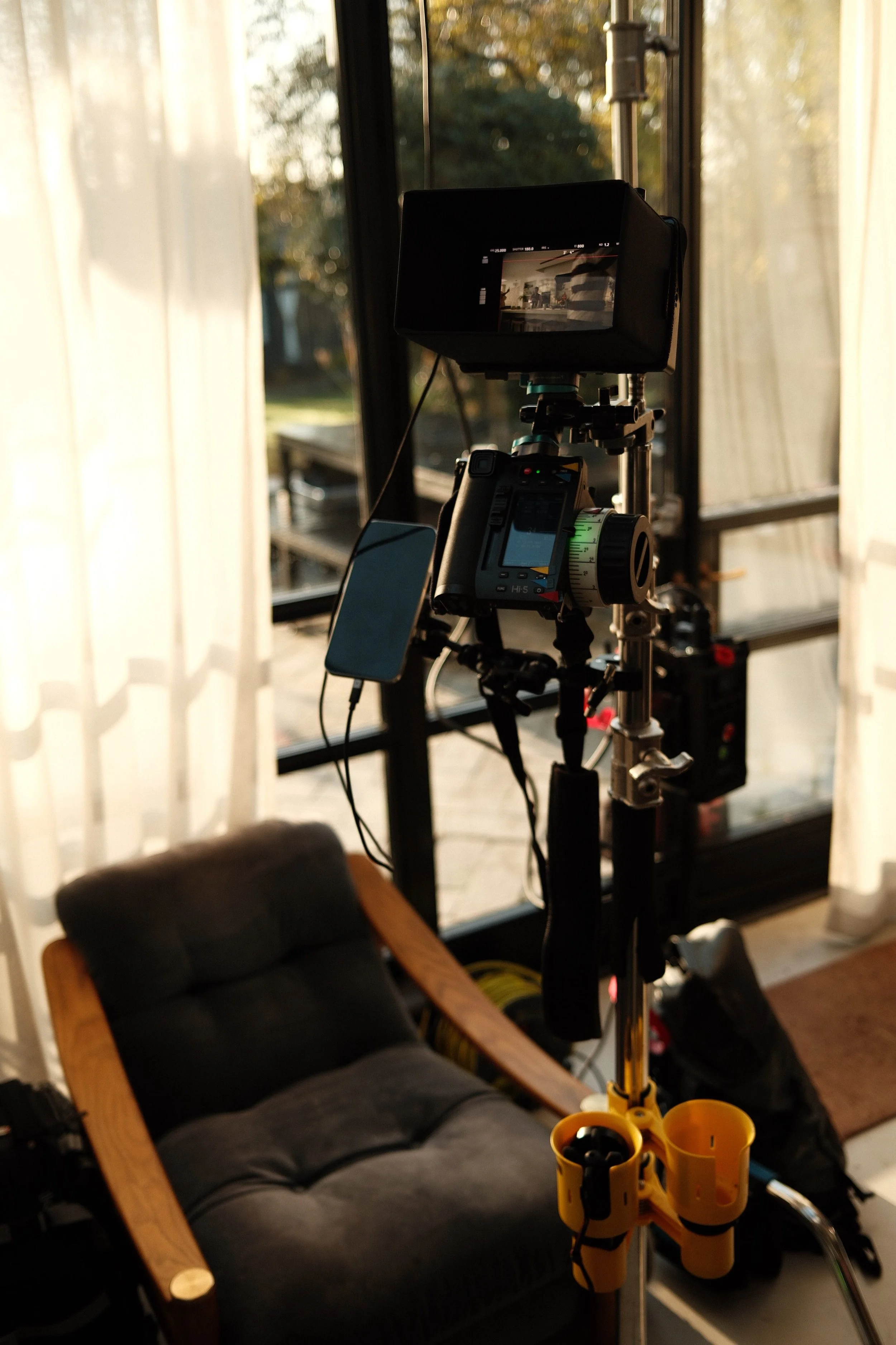 A camera setup with a monitor and phone on a tripod, in a room with large windows and sheer curtains, during sunset.