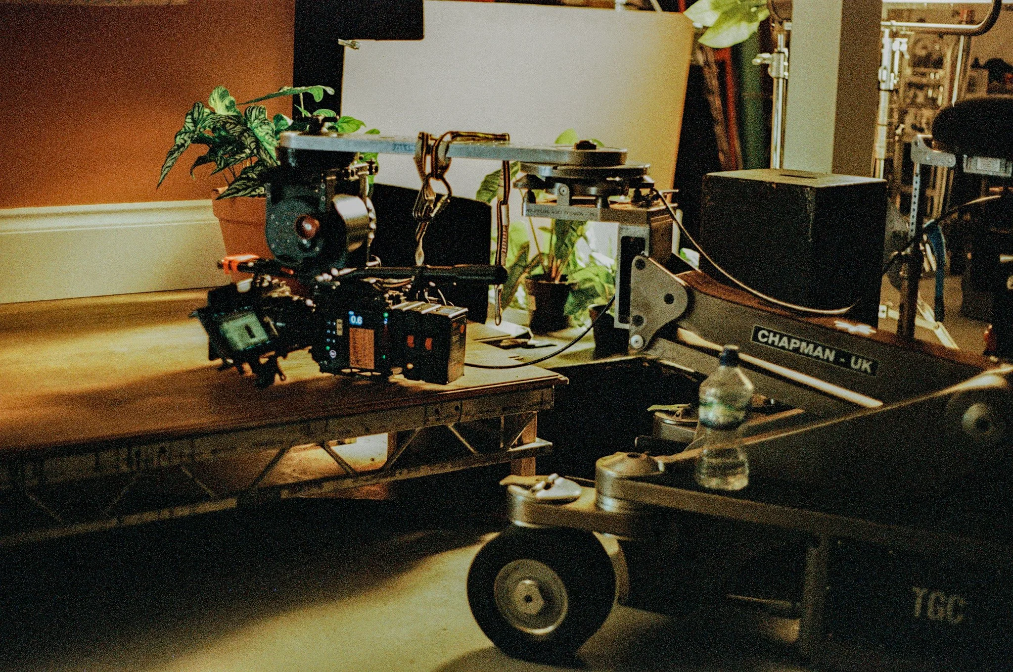 A photography setup with a camera mounted on a dolly or platform, surrounded by lighting equipment, monitors, and potted plants in a studio or shooting environment.