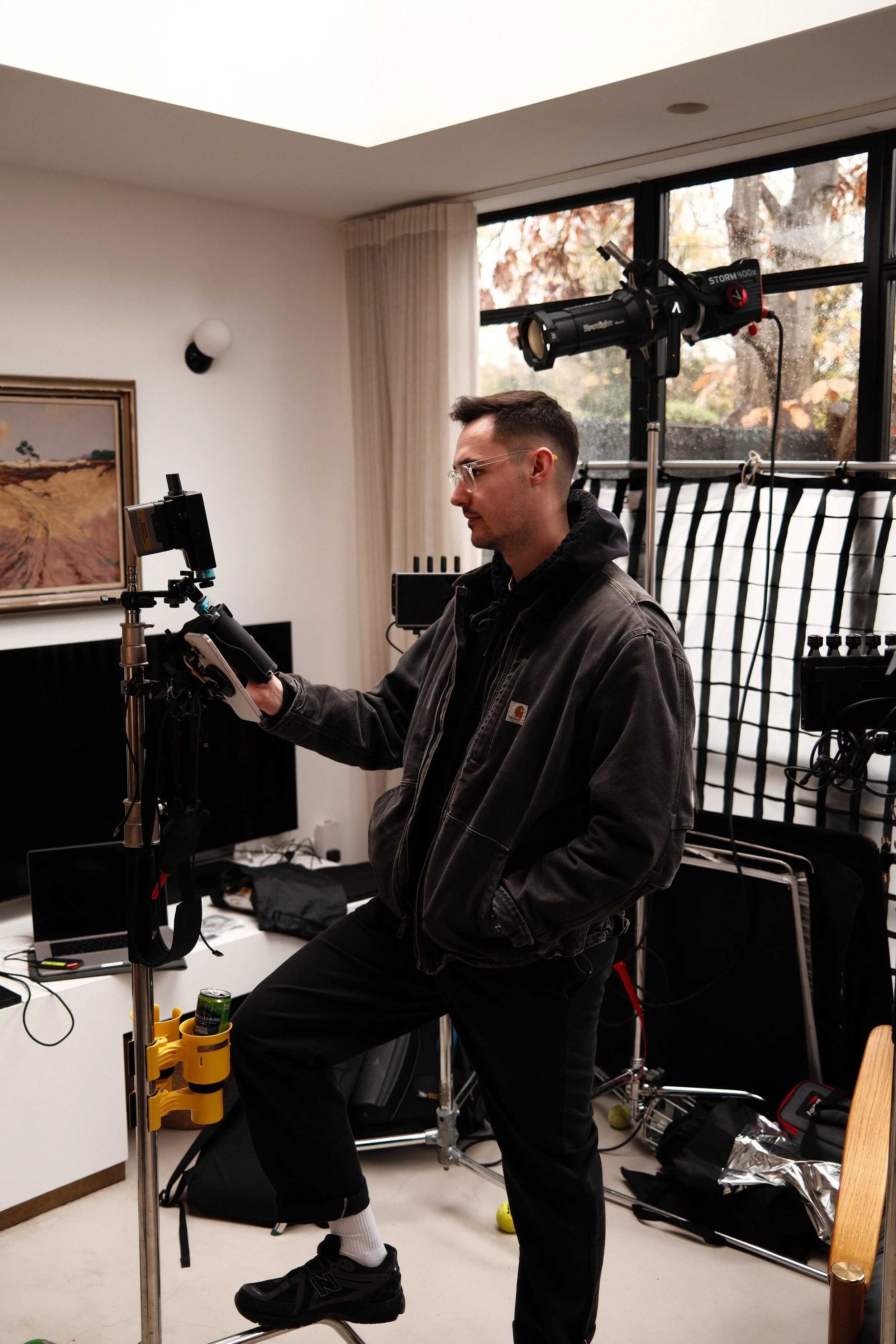 A man operating a camera rig in a room with a window showing trees outside.