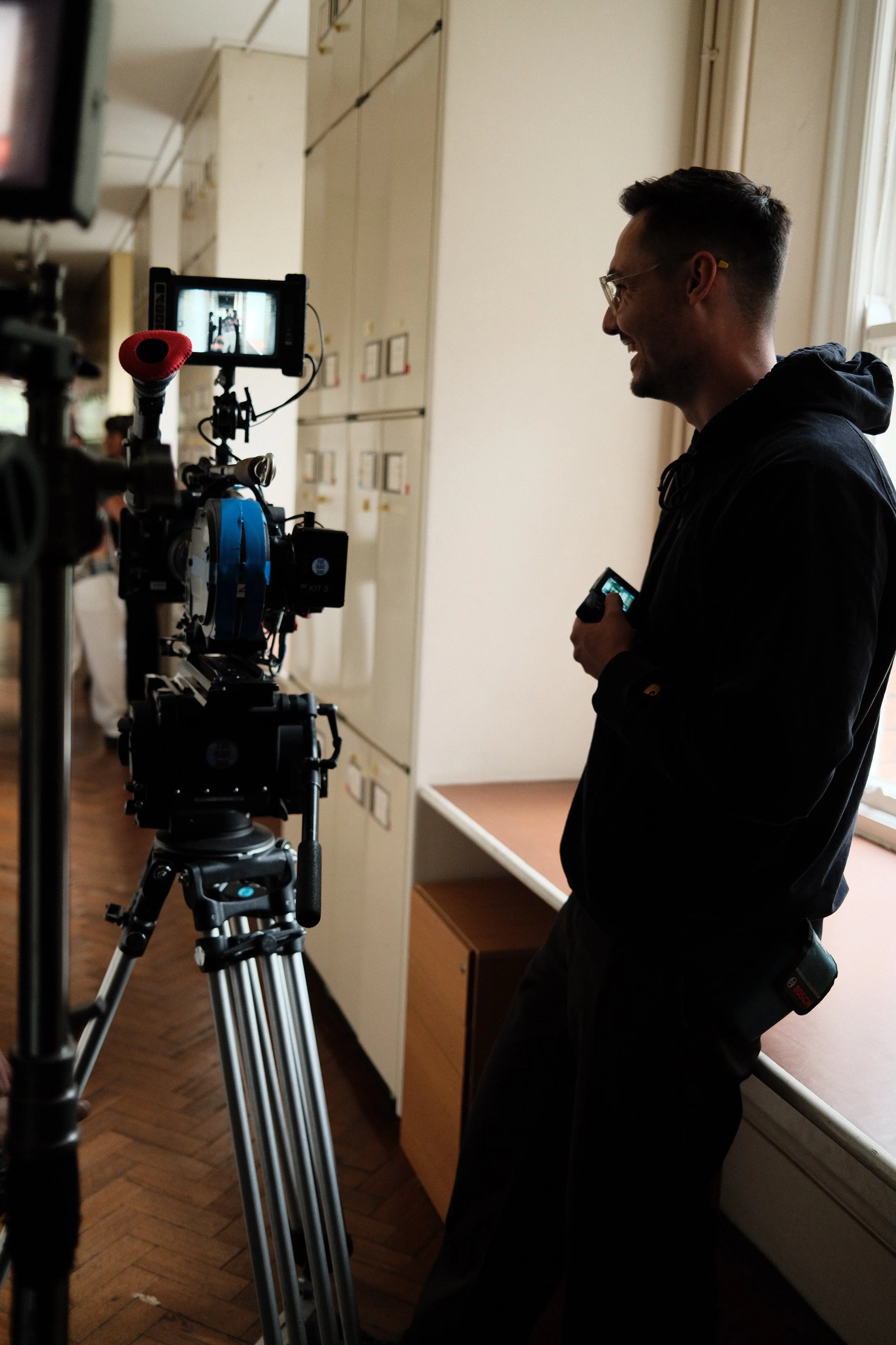 A man with glasses and a dark hoodie operates a professional camera on a tripod in a room with lockers, near a window with curtains.