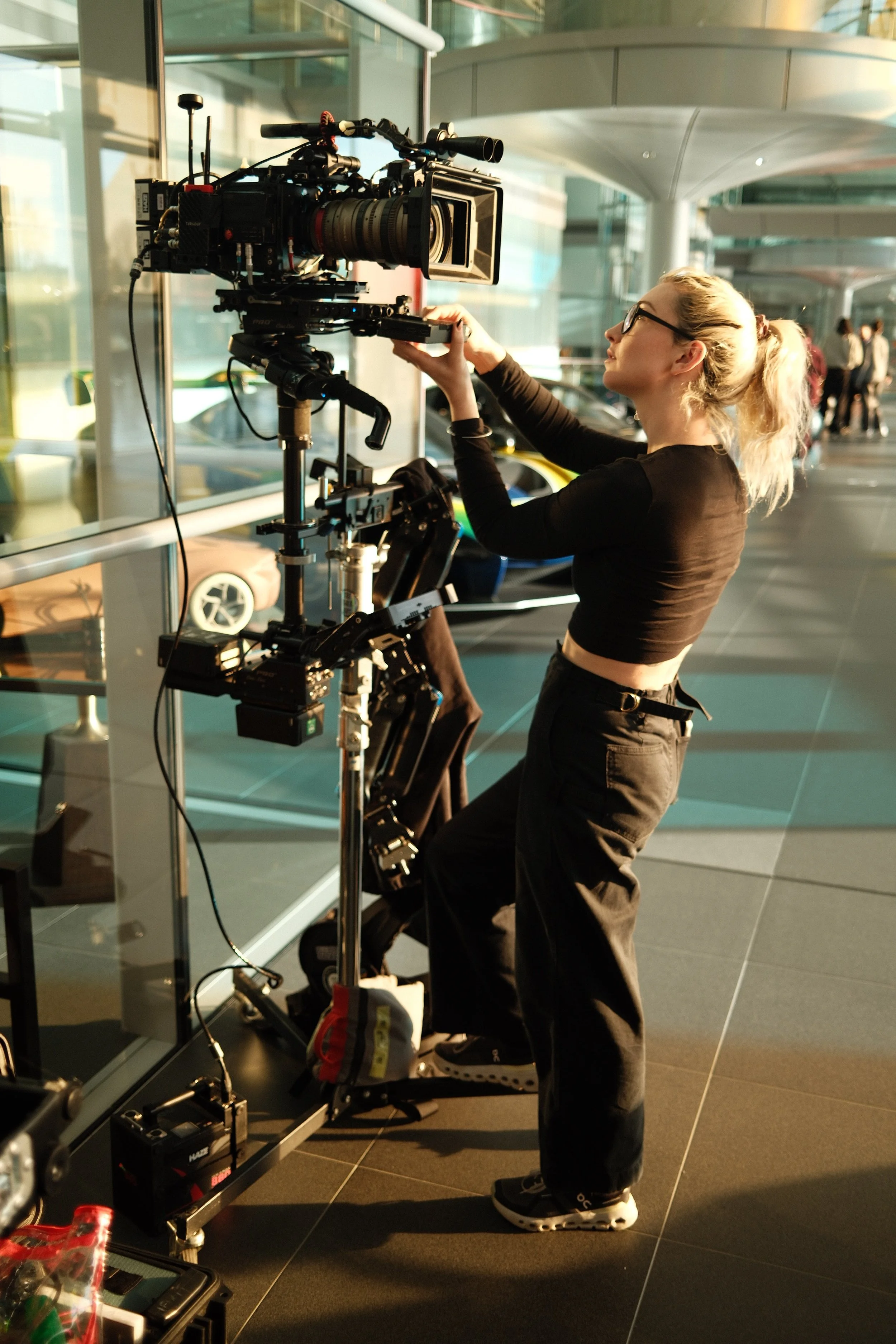 A woman operating a professional camera on a tripod inside a modern building with glass walls and a view of parked cars outside.