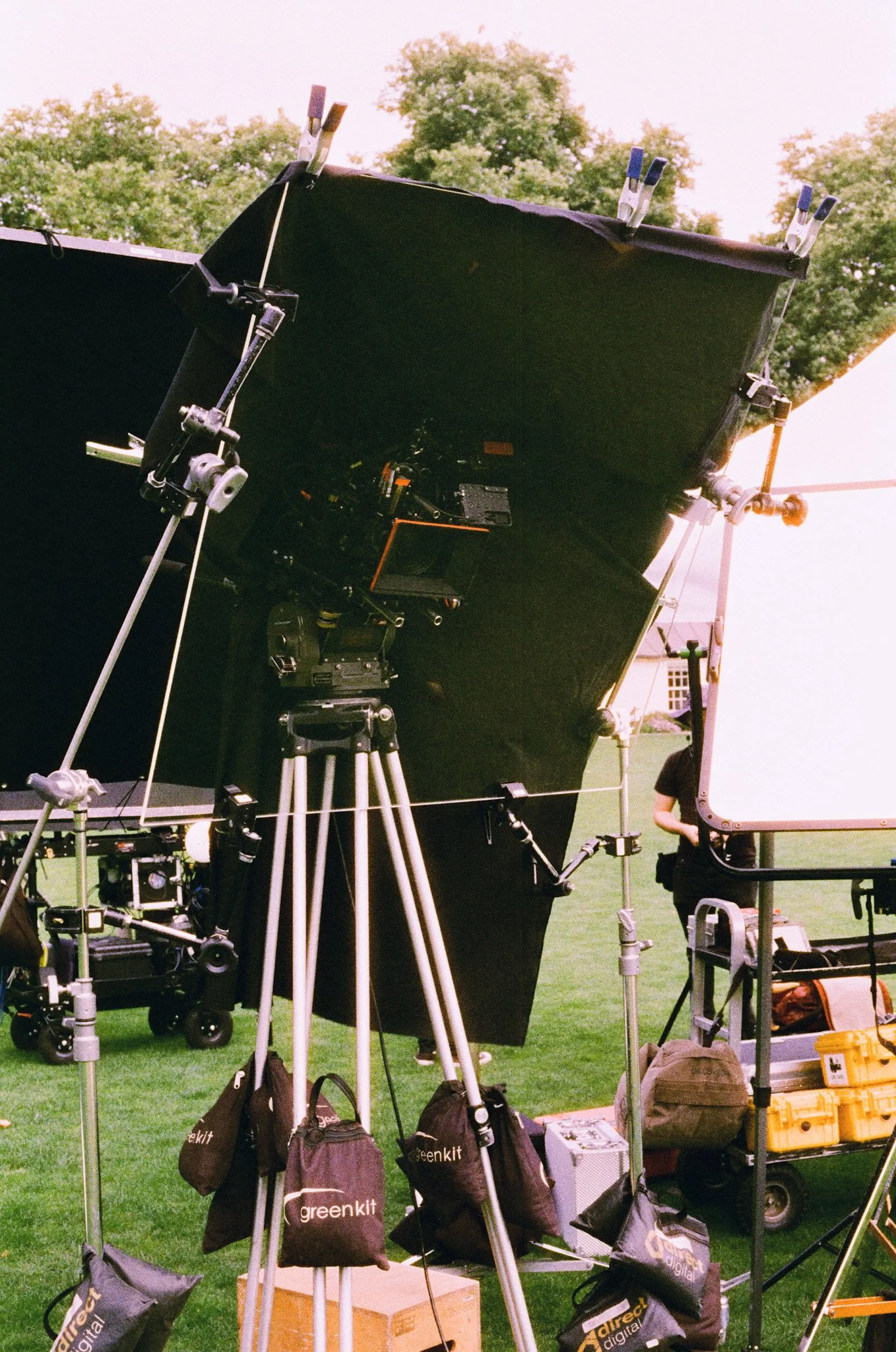 Film or photography shoot setup outdoors with cameras, tripods, lighting equipment, and bags on a grassy field, trees in the background.