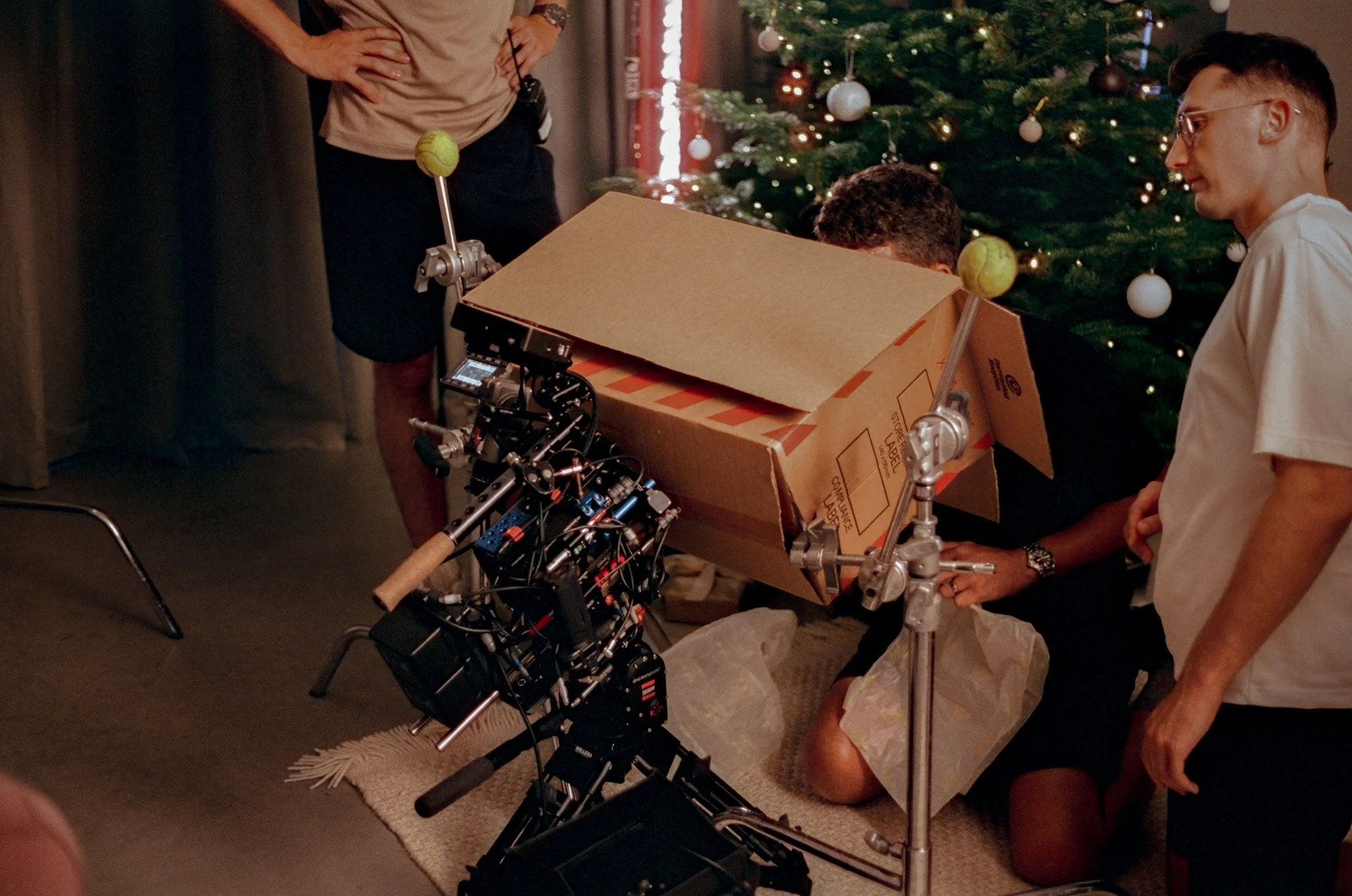 Filming crew building a Christmas scene with a camera, cardboard box, and Christmas tree in the background.