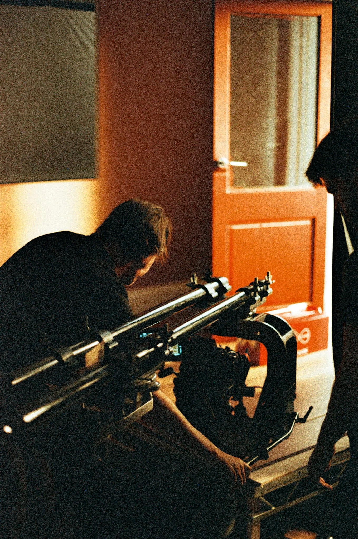 Two people adjusting camera equipment in a dimly lit room with warm lighting and a red door.