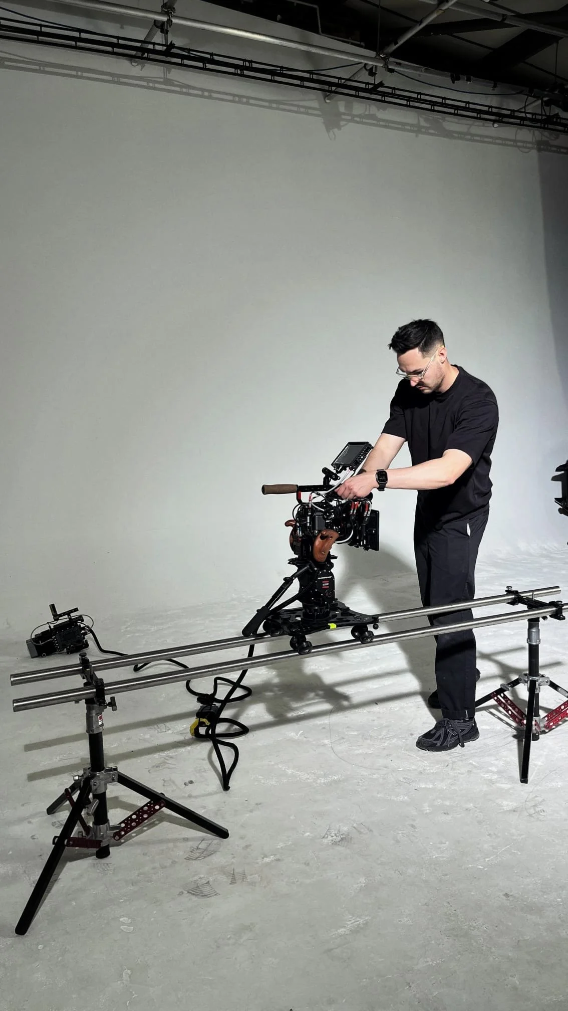 A man wearing black clothing and glasses is adjusting a professional camera mounted on a slider in a studio with a blank white wall and ceiling pipes.
