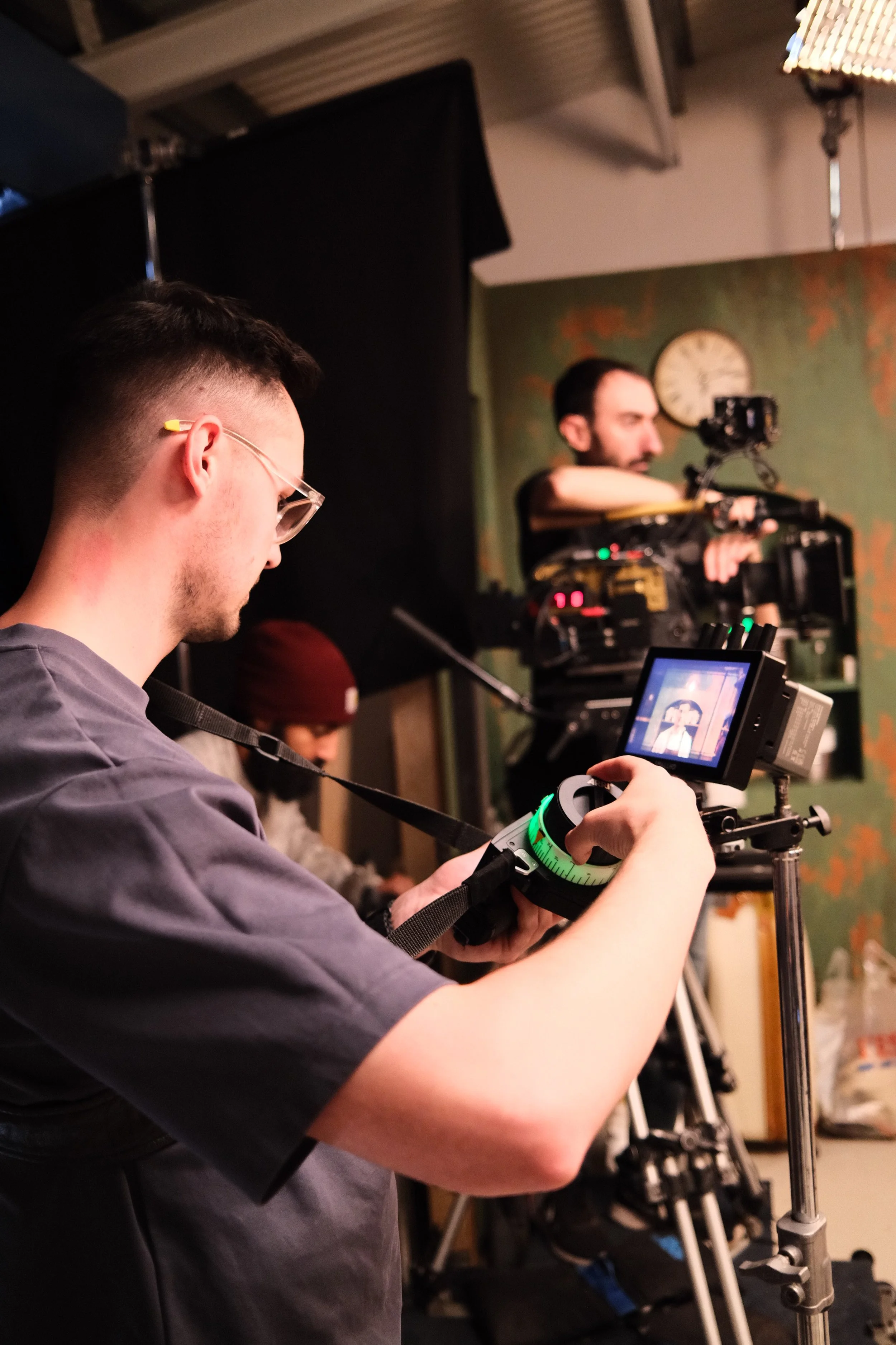 A person operating a camera with a monitor in a filming or photography studio. In the background, another person is working with equipment, and a cameraman is filming with a professional camera on a tripod. A clock and a patterned wall are visible.