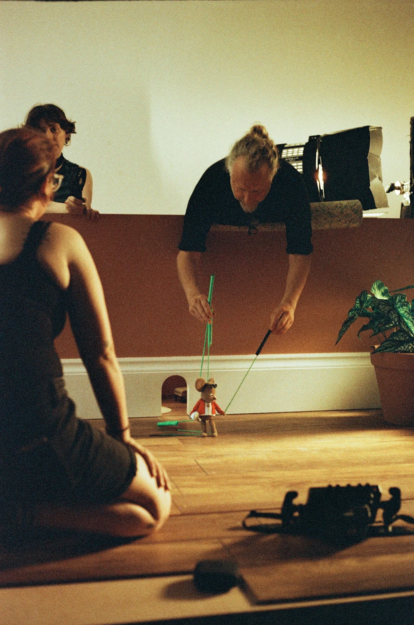 A person leans over to adjust a marionette puppet resembling a bear, while two women watch. The scene appears to be indoors with a large plant on the right and a dark object in the foreground.