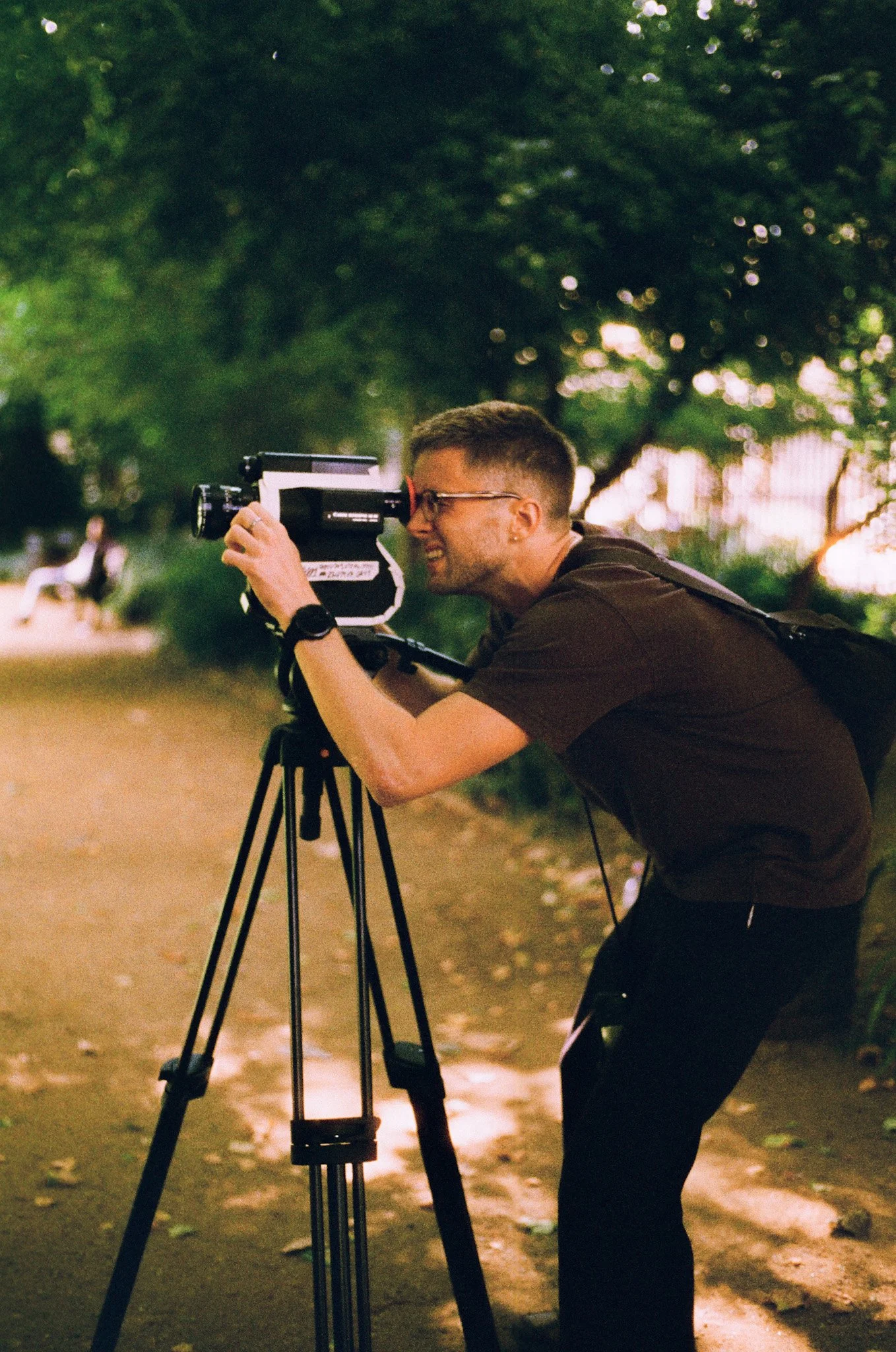 A man with glasses, wearing a black shirt, is using a large camera mounted on a tripod outdoors. He is leaning forward, looking through the camera's viewfinder, with trees and a woman sitting on a bench blurred in the background.