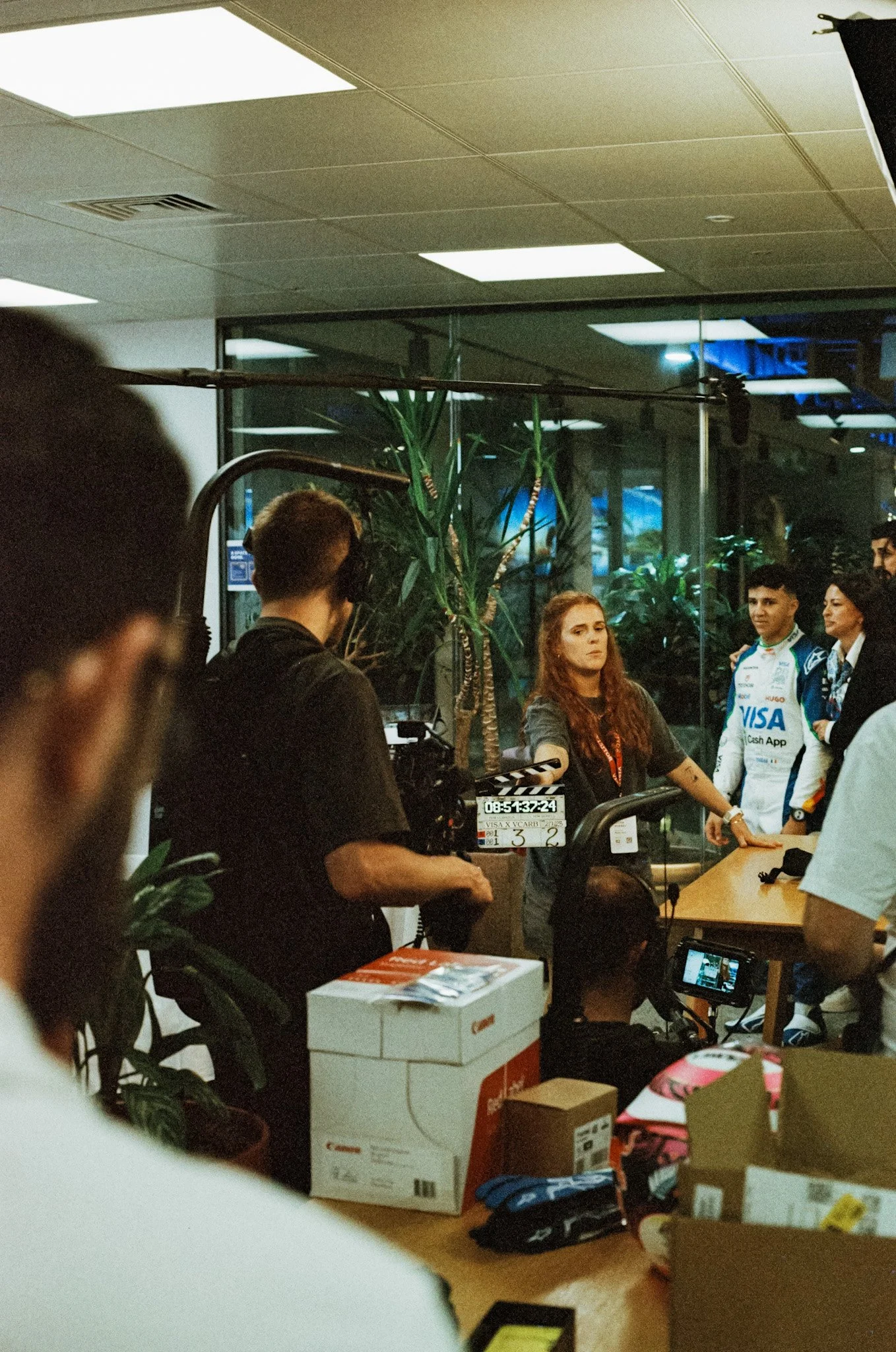 Filming crew filming a woman at an indoor interview or press event with people in racing suits in the background.