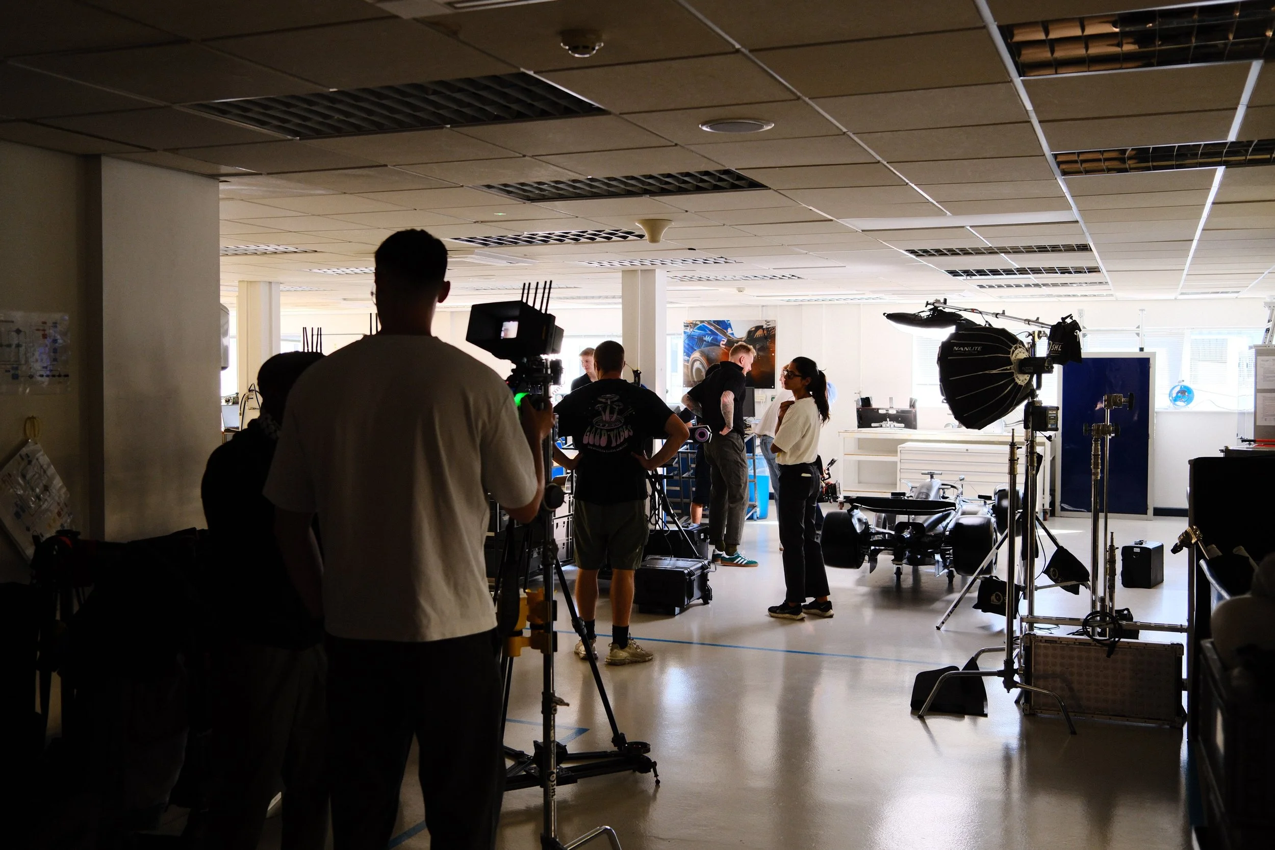 A film crew on set in an indoor studio, with various professional lighting and camera equipment, and a woman in white shirt and black pants being filmed or interviewed.