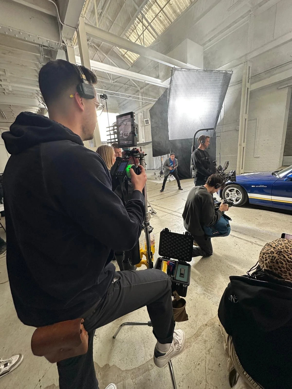 Behind the scenes of a film or photo shoot with a blue sports car, crew members operating cameras, lights, and equipment in a spacious industrial setting.