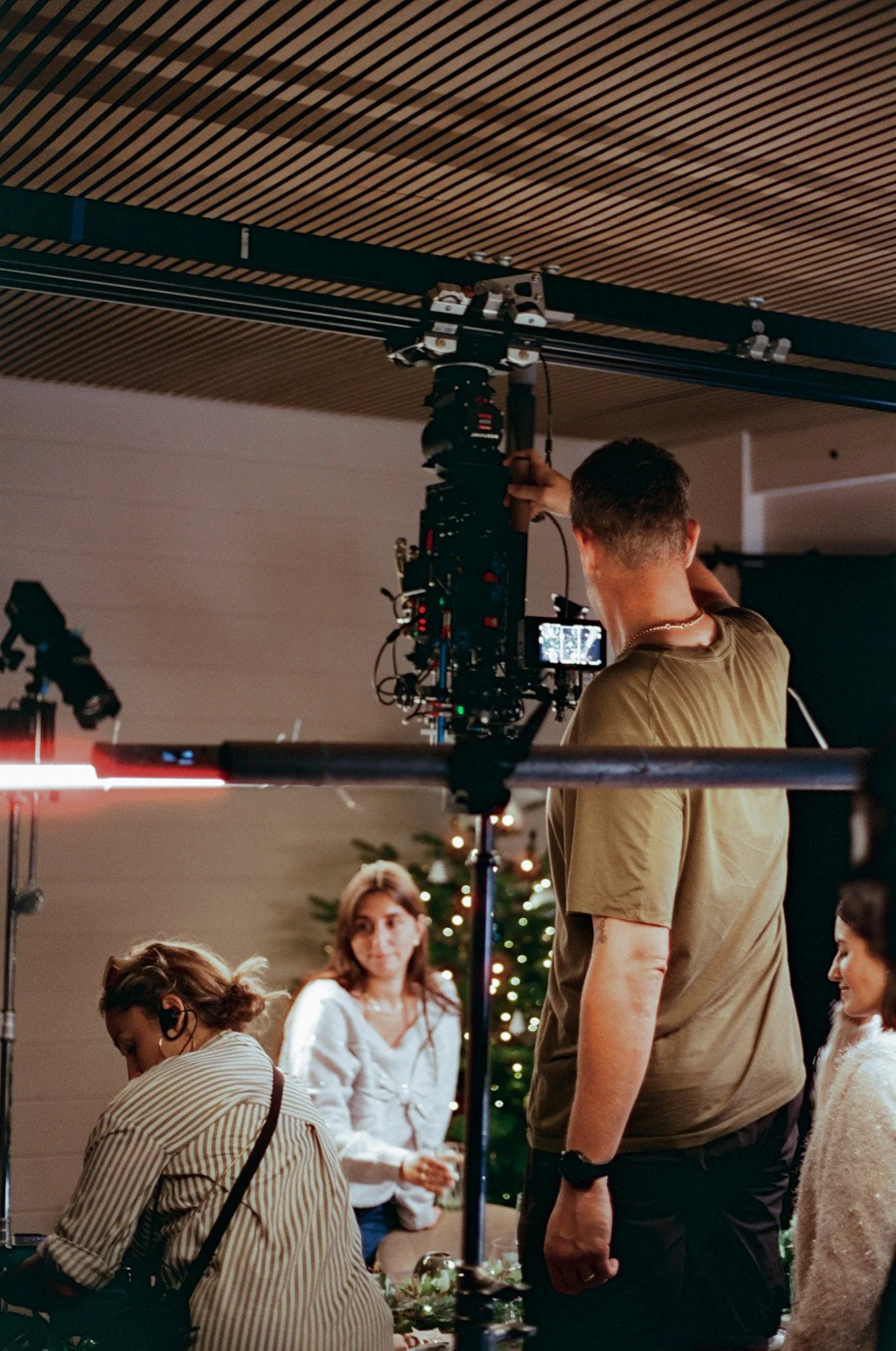 A behind-the-scenes shot of a film or photo shoot during a holiday celebration with a decorated Christmas tree in the background. Several people are working or preparing, including a woman in a striped shirt with earpiece, and a man operating camera 
