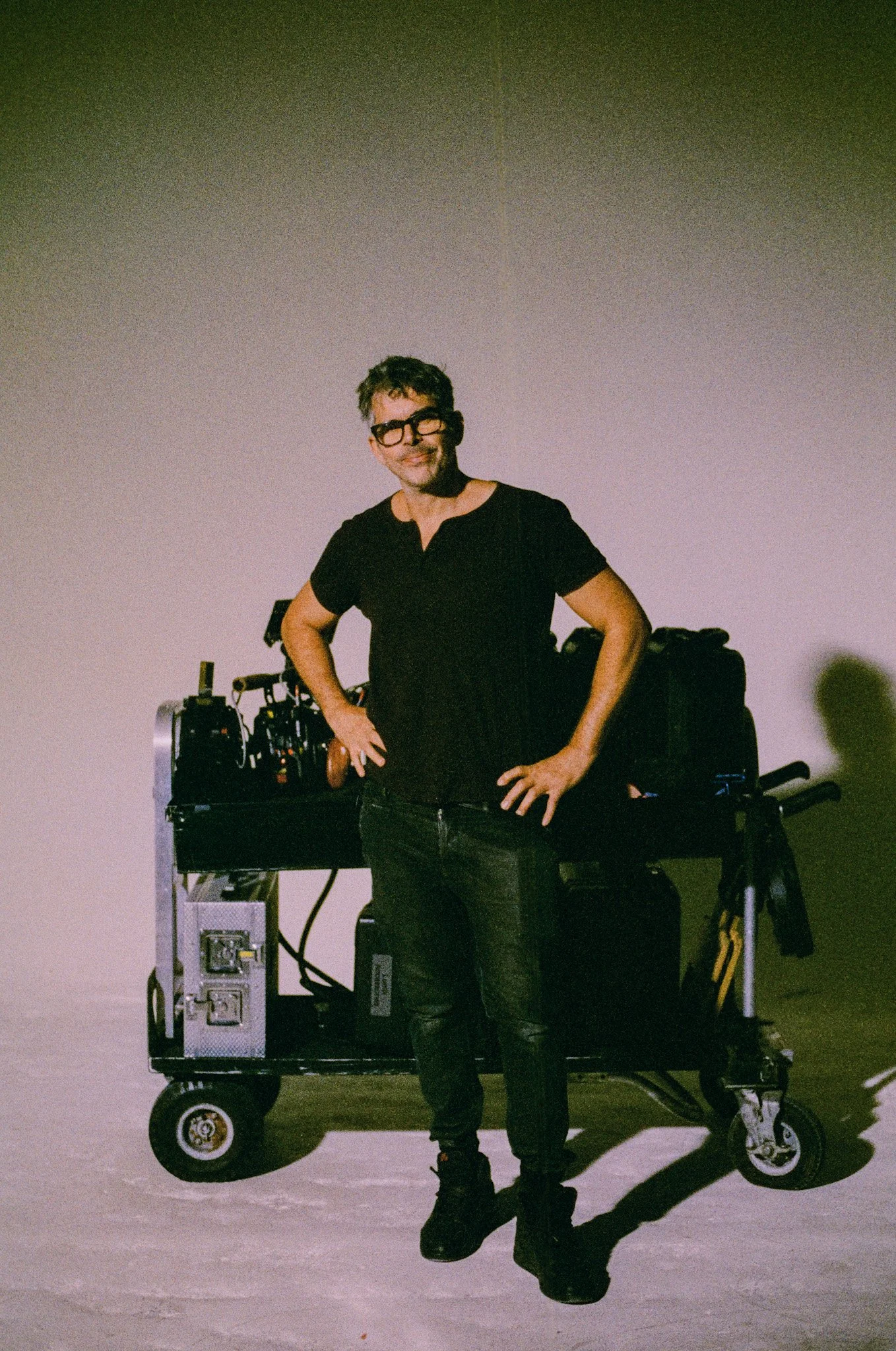A man with glasses, wearing a black shirt and jeans, stands with hands on hips in front of a cart filled with filming equipment in a large, empty space.