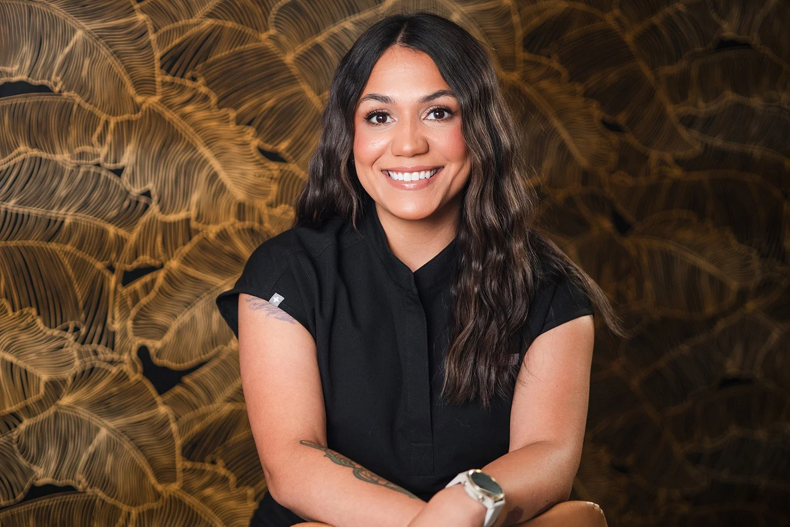Woman smiling, sitting on chair against black background, wearing black button-up shirt and gray jeans.