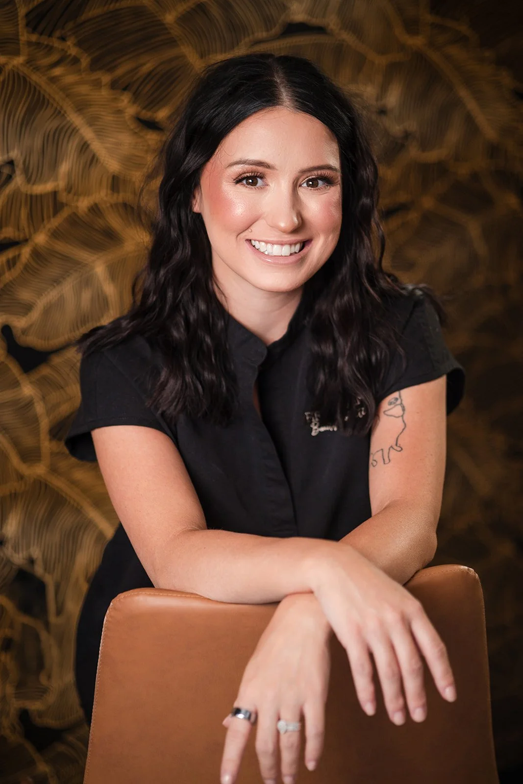 A woman with long, dark hair, smiling and seated on a beige chair. She is wearing a black top with sheer, pearl-studded sleeves. The background is black.