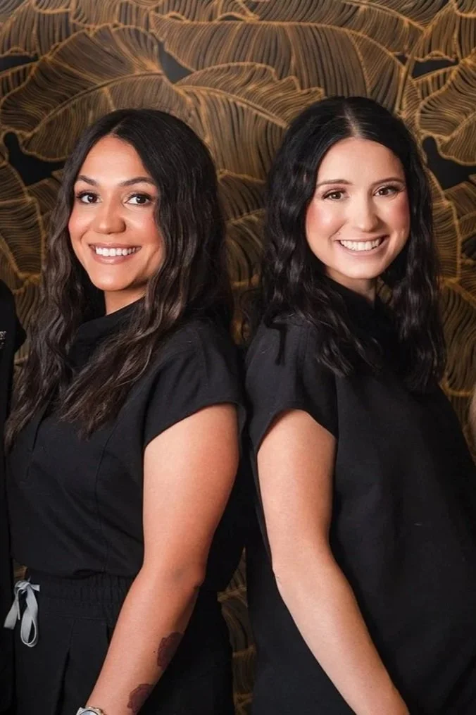 Two women smiling, standing back to back against a dark background. Both wear black tops and blue jeans.