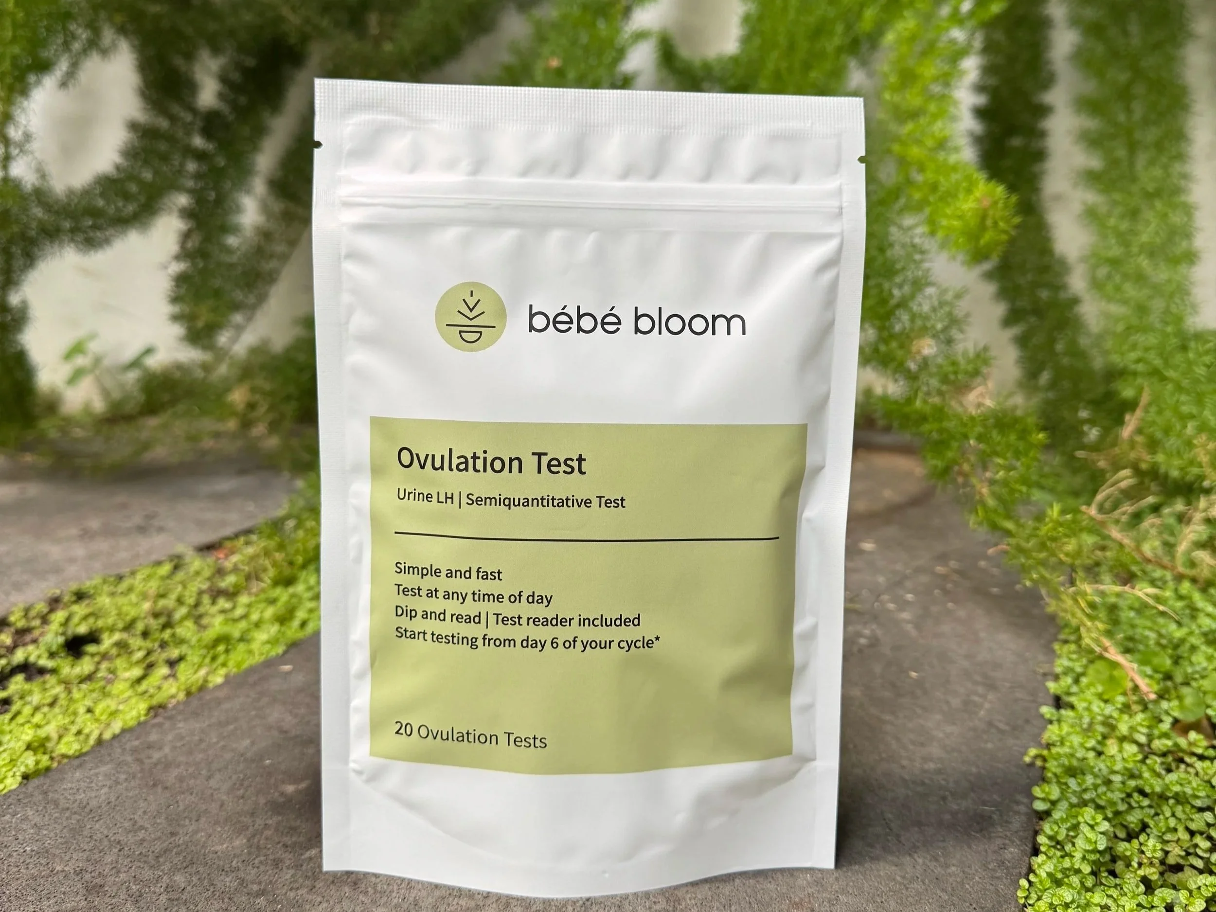 bébé bloom Ovulation Test pouch displayed outdoors against lush green foliage, showing product label with "Urine LH | Semiquantitative Test" and 20 Ovulation Tests.