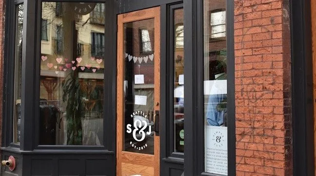 Storefront with a wooden door and large glass windows, decorated with white and pink paper hearts, and a circular sign with the number 8 and the word 'Seattle'.