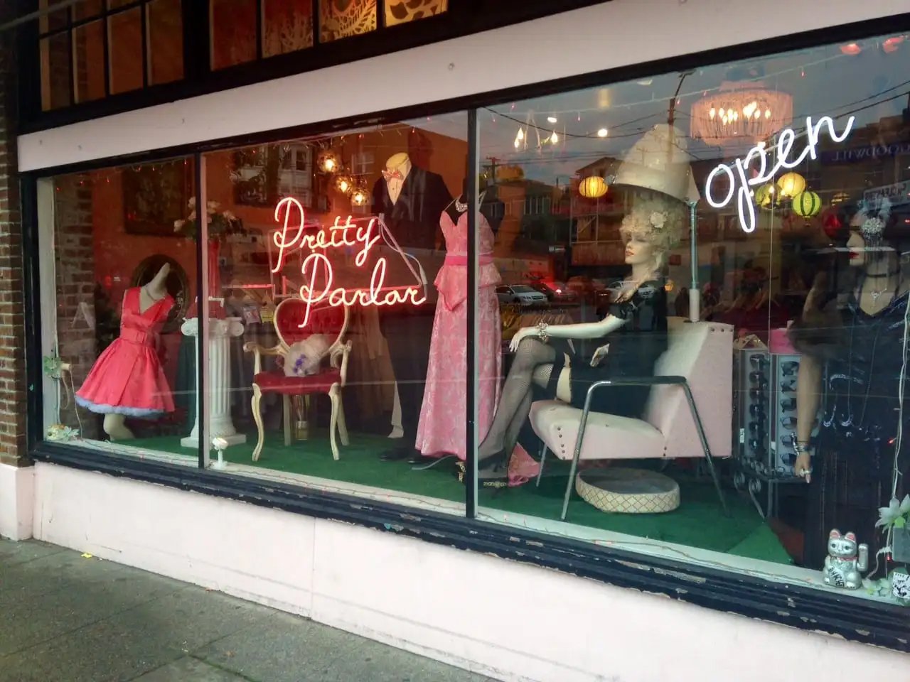 Clothing boutique window display with mannequins wearing vintage-style dresses, neon signs reading 'Pretty Parlour' and 'Open,' and decorative items like a stuffed animal and a large hat.