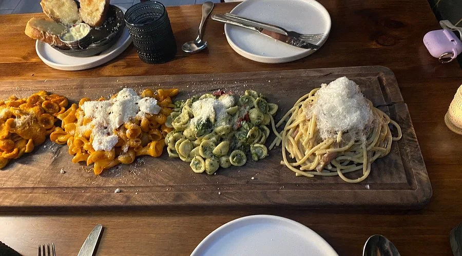 Four types of pasta on a wooden board: yellow tortellini, yellow pasta with white cheese, green pasta with vegetables, and spaghetti with white cheese.