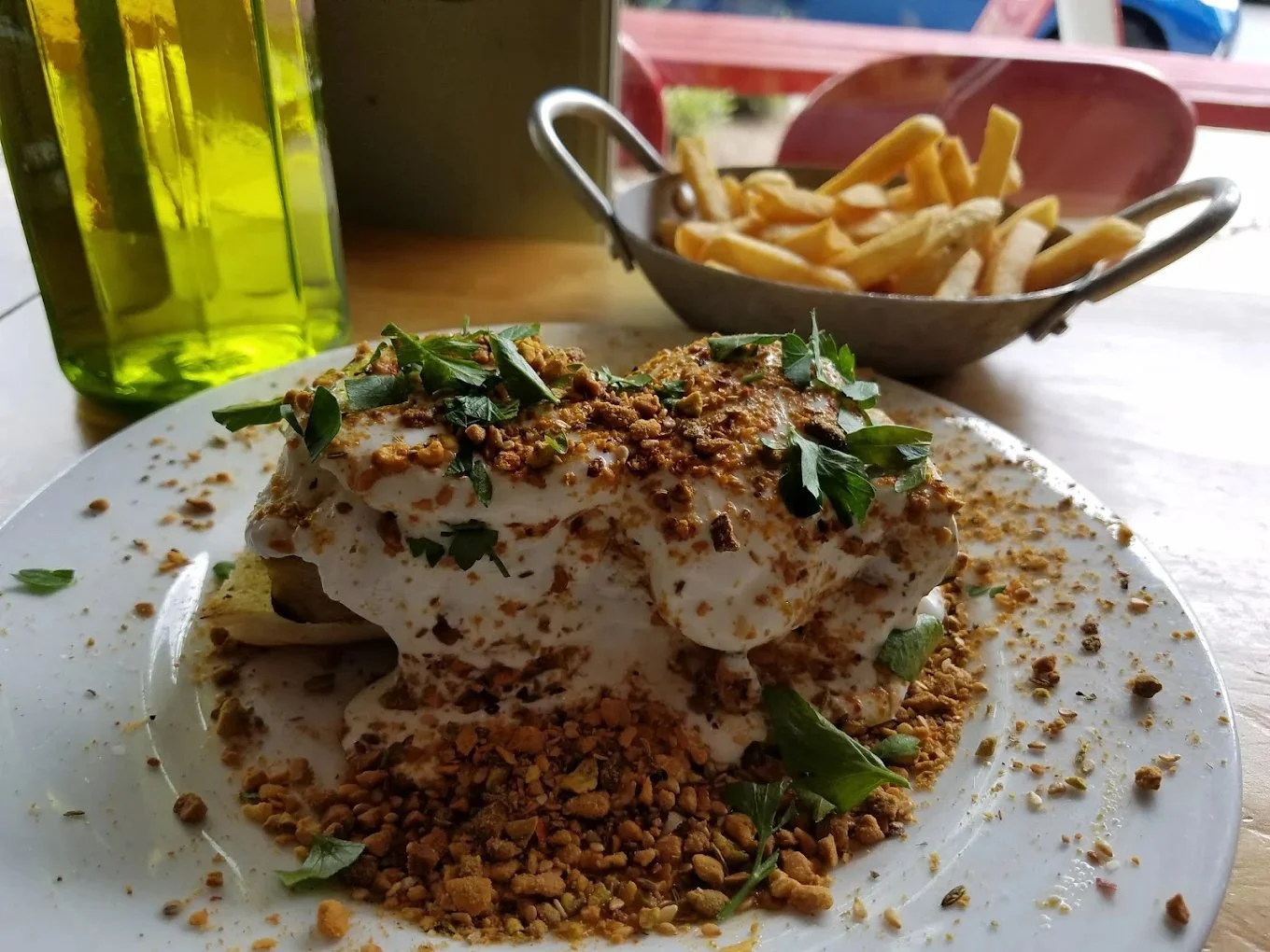 A plate of food with a fried egg topped with chopped herbs and crushed nuts, surrounded by more crushed nuts on a white plate. In the background, a bowl of French fries and a tall green bottle are visible, set on a wooden table with a blurred outdoor setting.