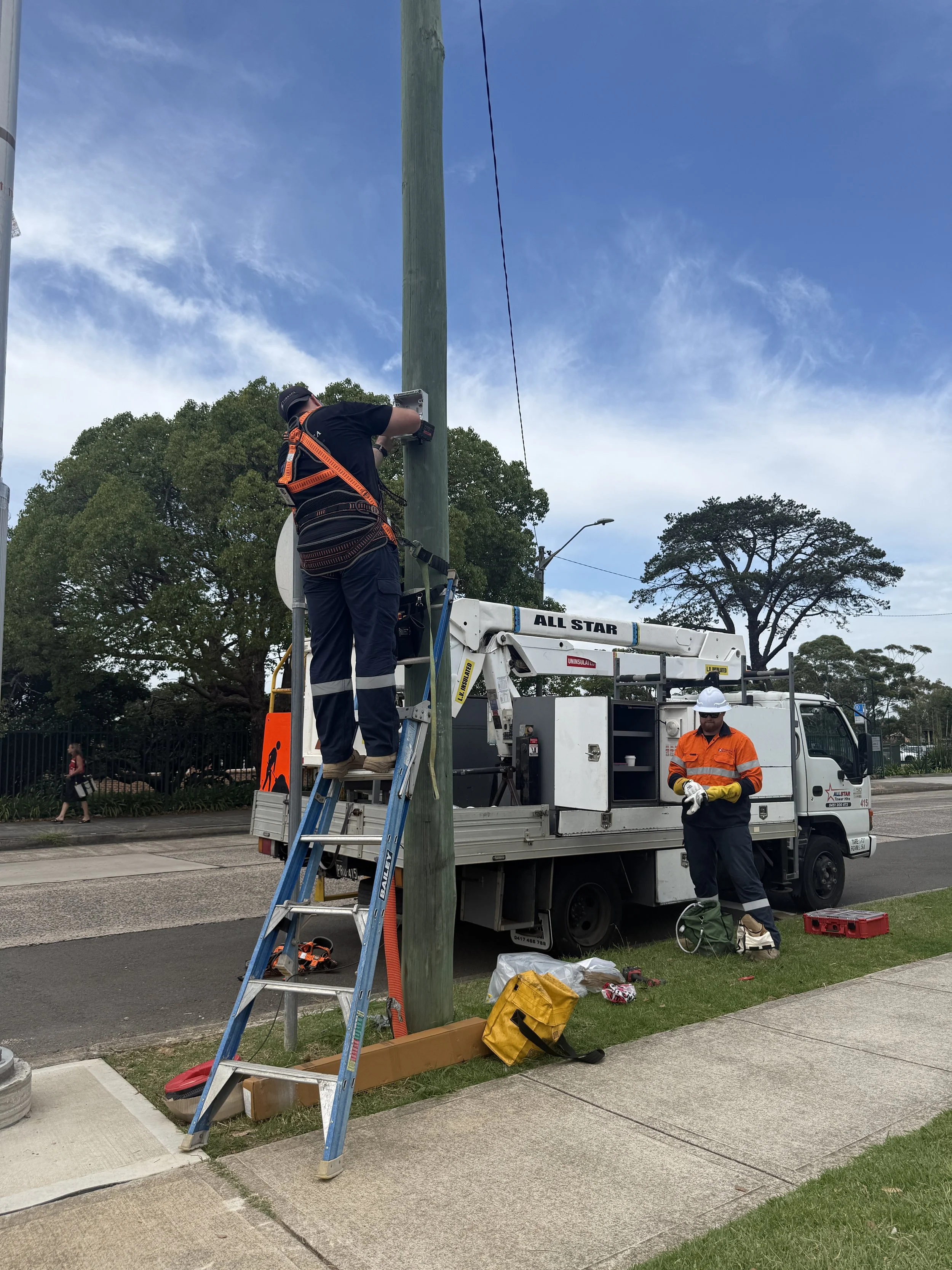 Two utility workers repairing a streetlight pole with a work truck and ladder nearby.