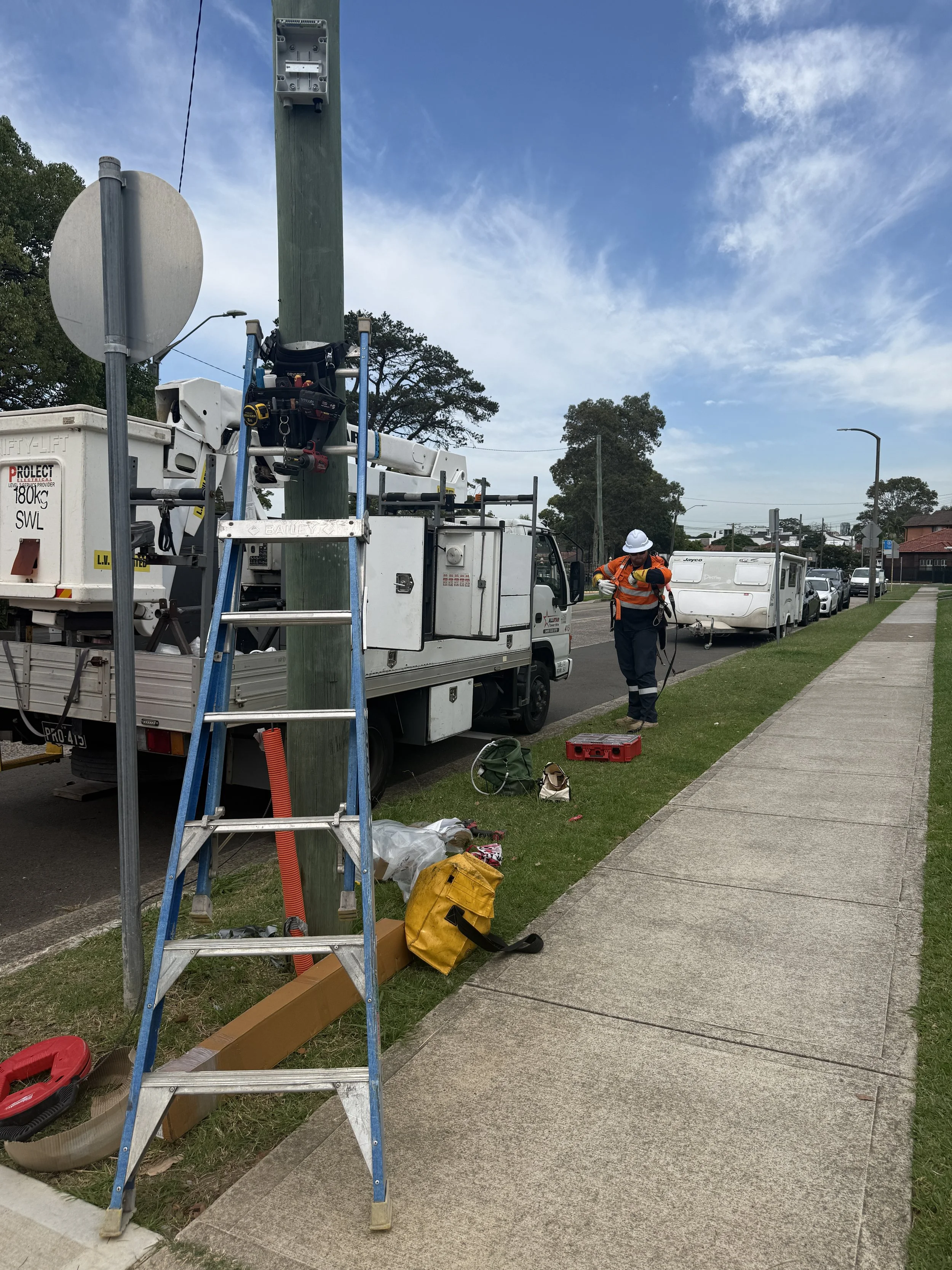 Worker repairing electrical or utility pole on roadside with equipment and tools, adjacent to a sidewalk and parked vehicles.