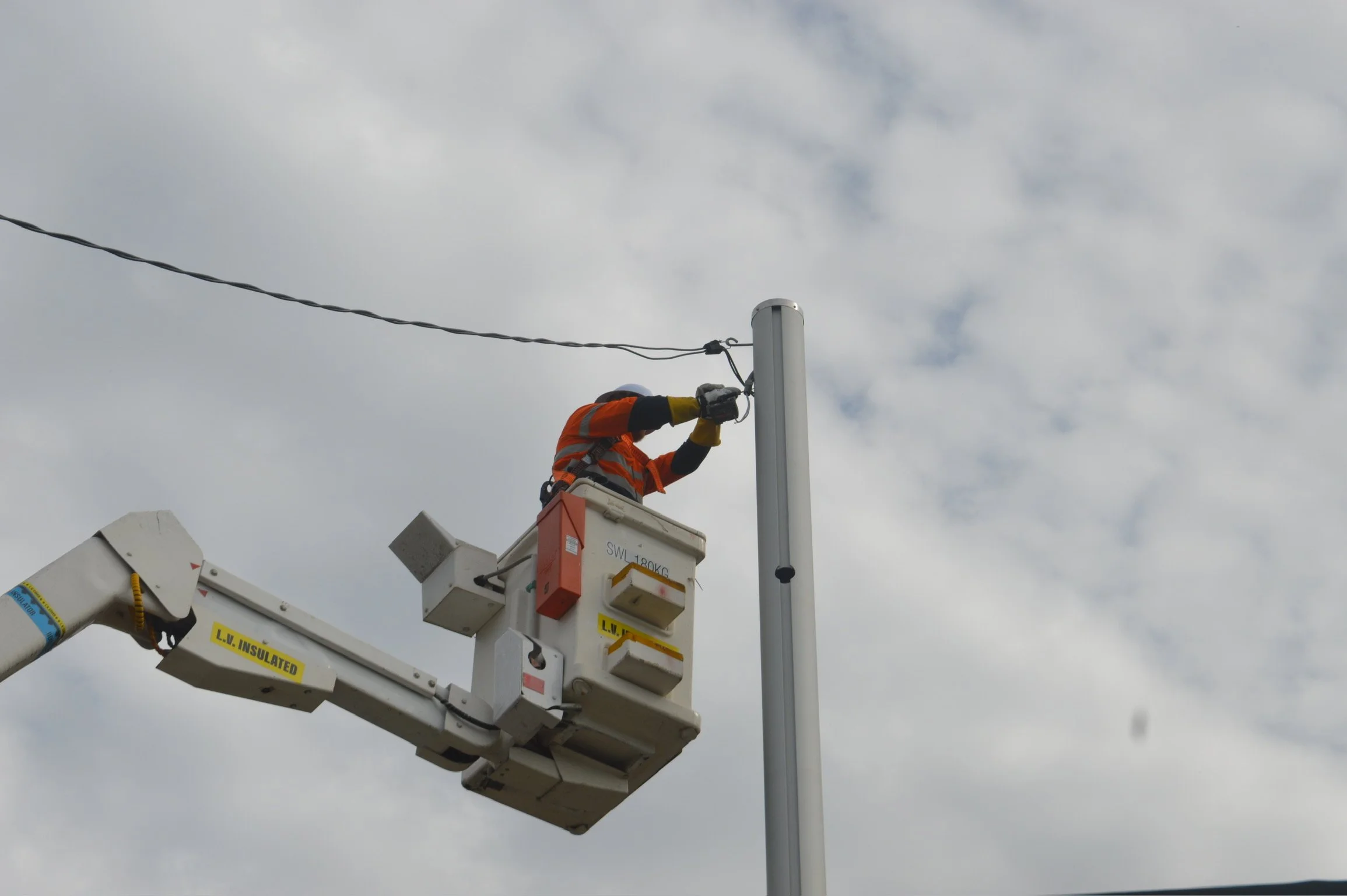 Worker in an orange safety vest and helmet standing in a bucket lift, installing or repairing a utility pole against a cloudy sky.