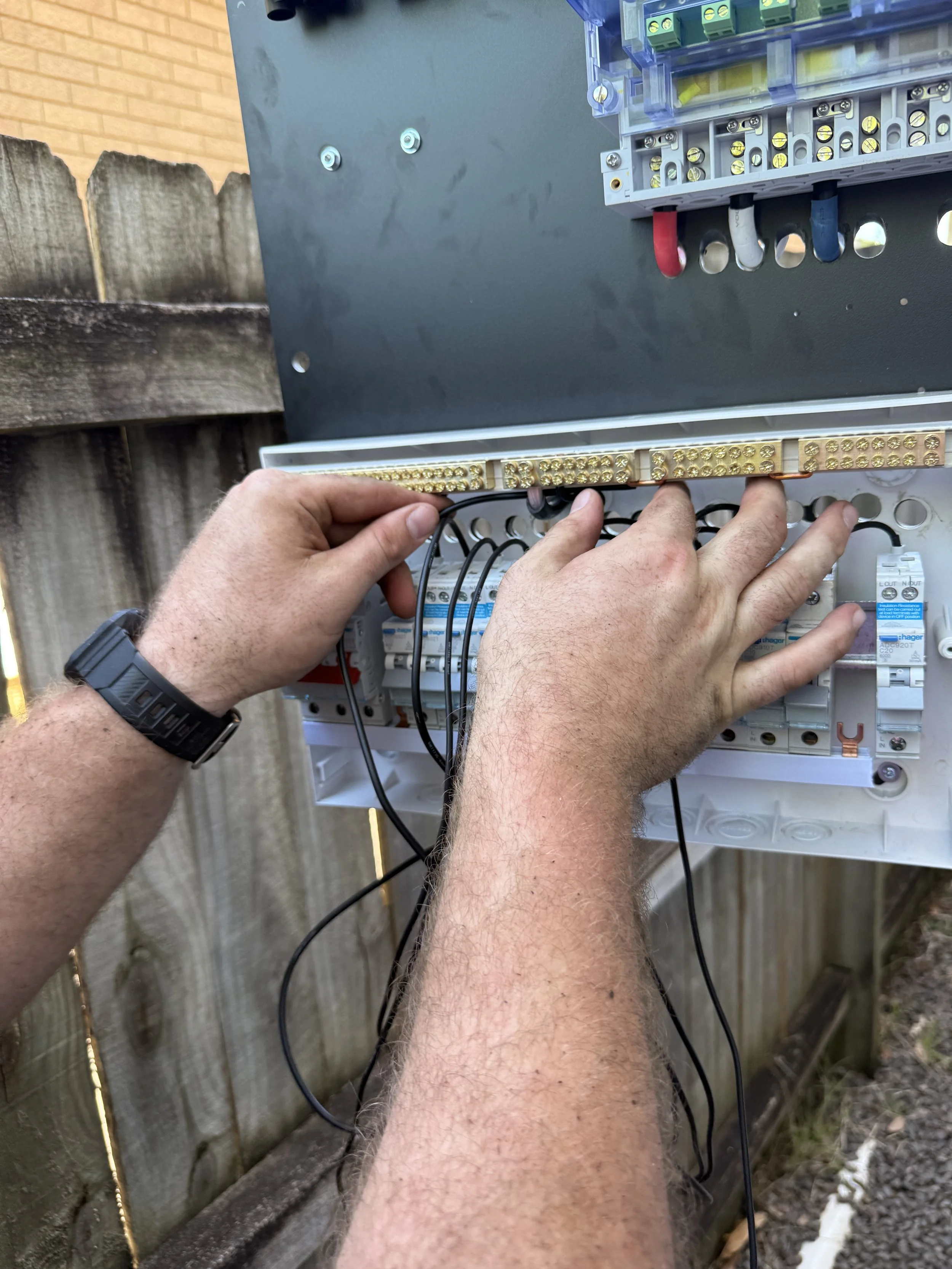 Person working on electrical wiring in an outdoor breaker box mounted on a wooden fence.