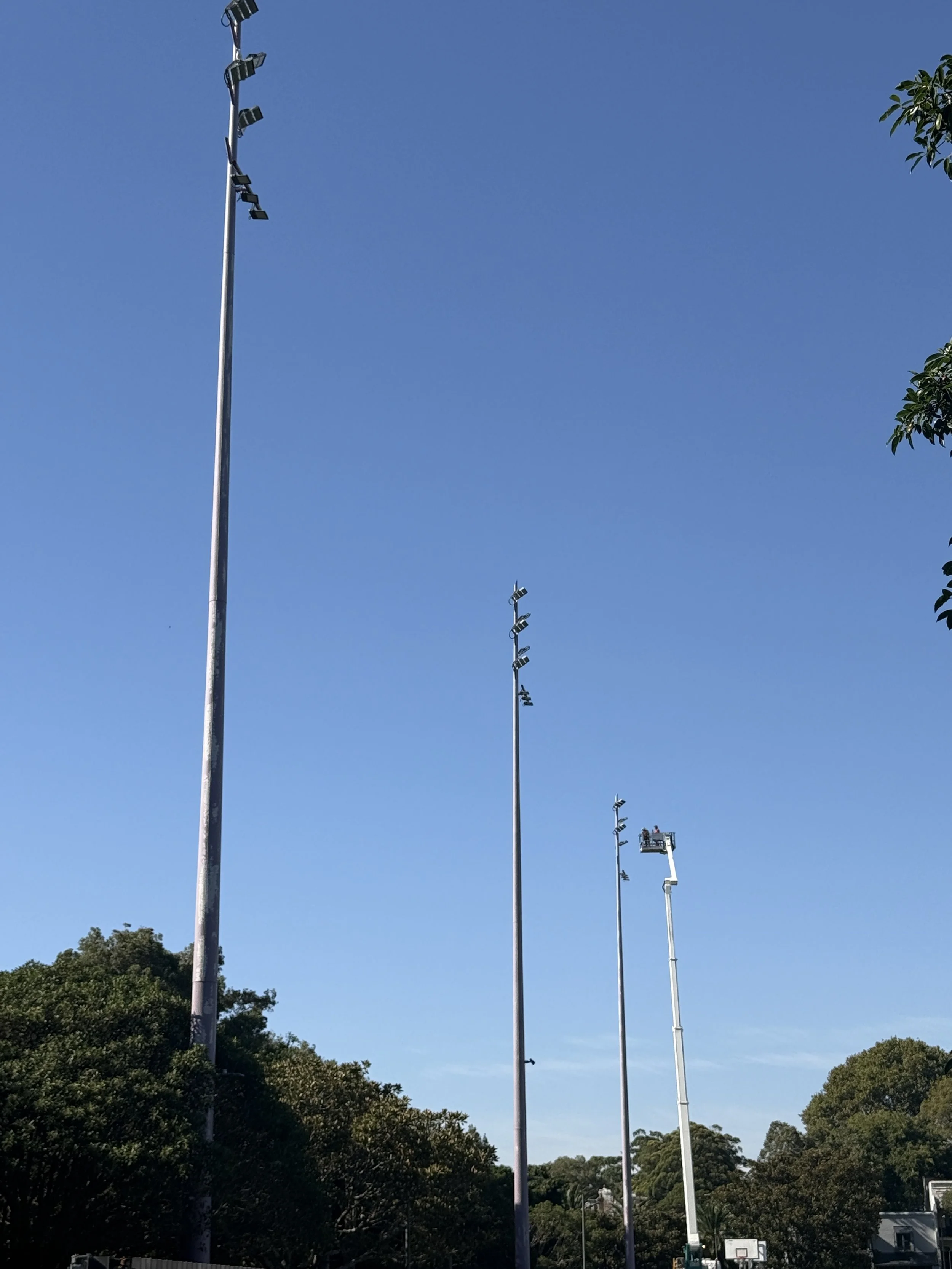 Four tall light poles on a sports field, with trees and a basketball hoop in the background, under a clear blue sky.