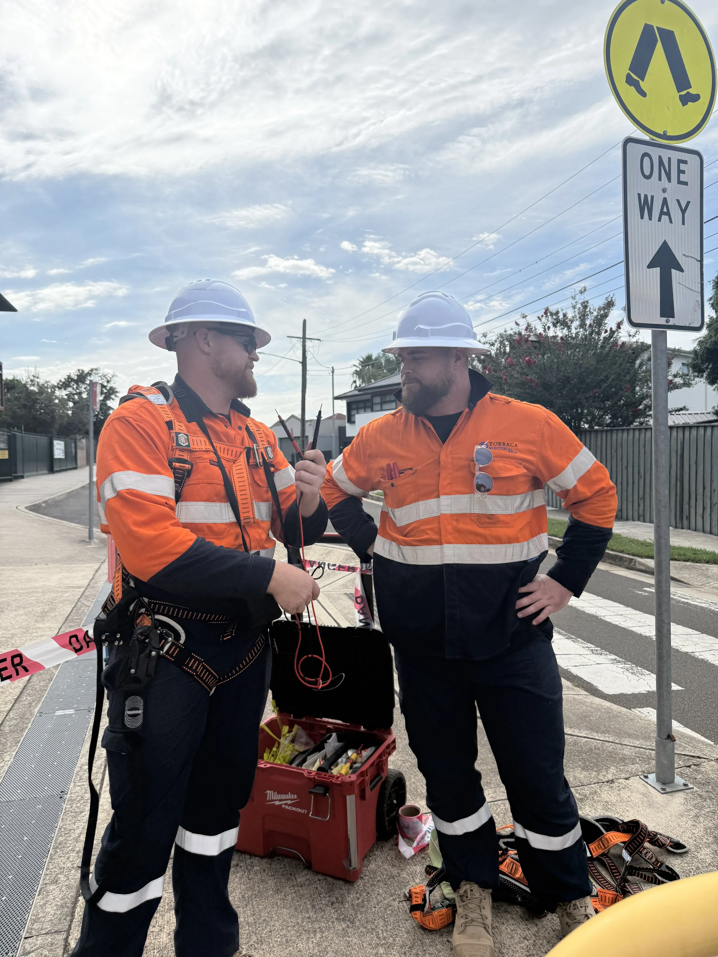 Two utility workers in orange safety uniforms and white helmets standing on a sidewalk, having a conversation. One worker is holding a walkie-talkie. There is a toolbox and safety equipment on the ground nearby, with traffic and street signs in the b