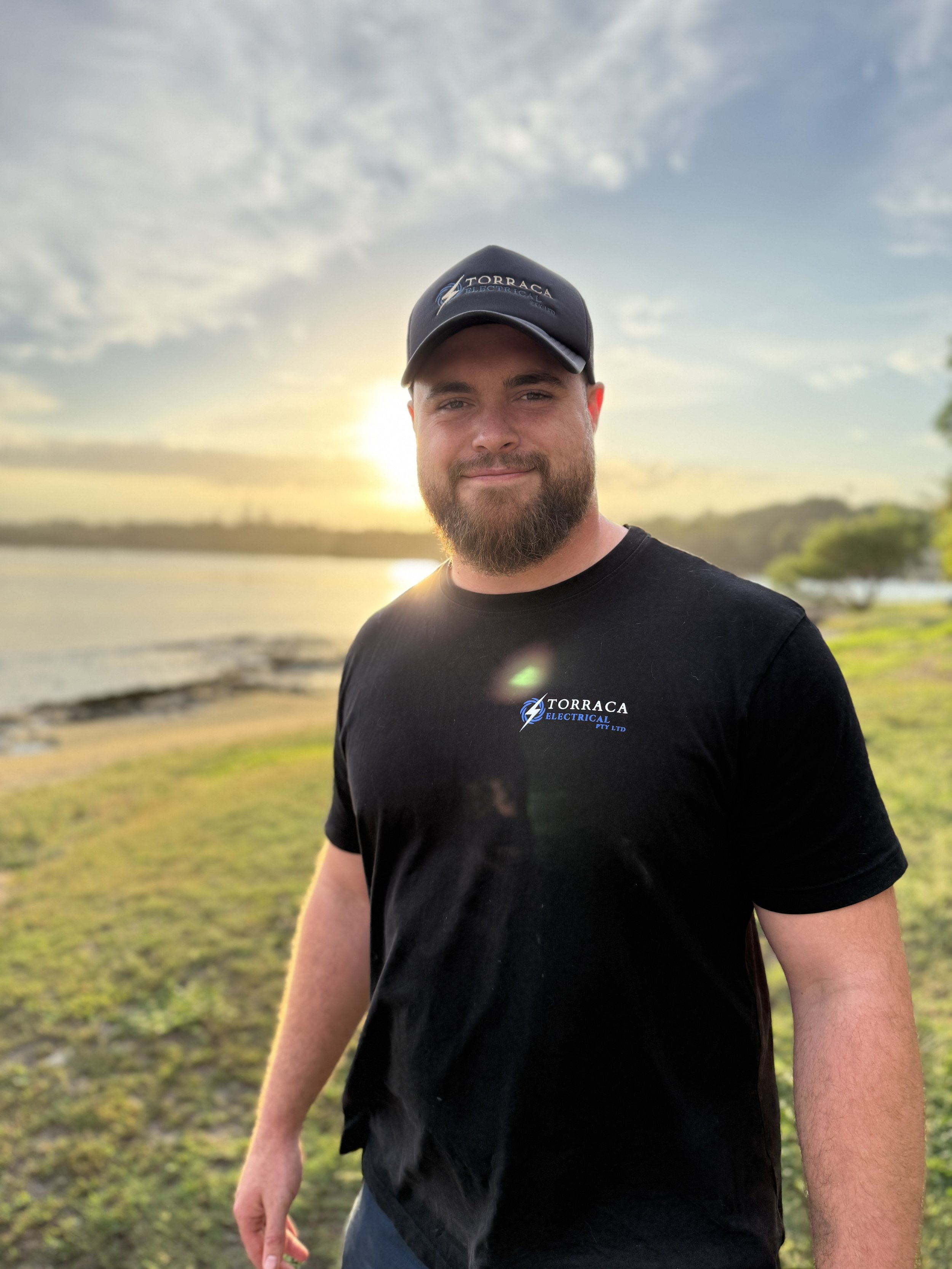 A man with a beard smiling at the camera outdoors during sunset, wearing a black T-shirt and cap with the logo PT Liquor Store logo and Torraca Electrical.