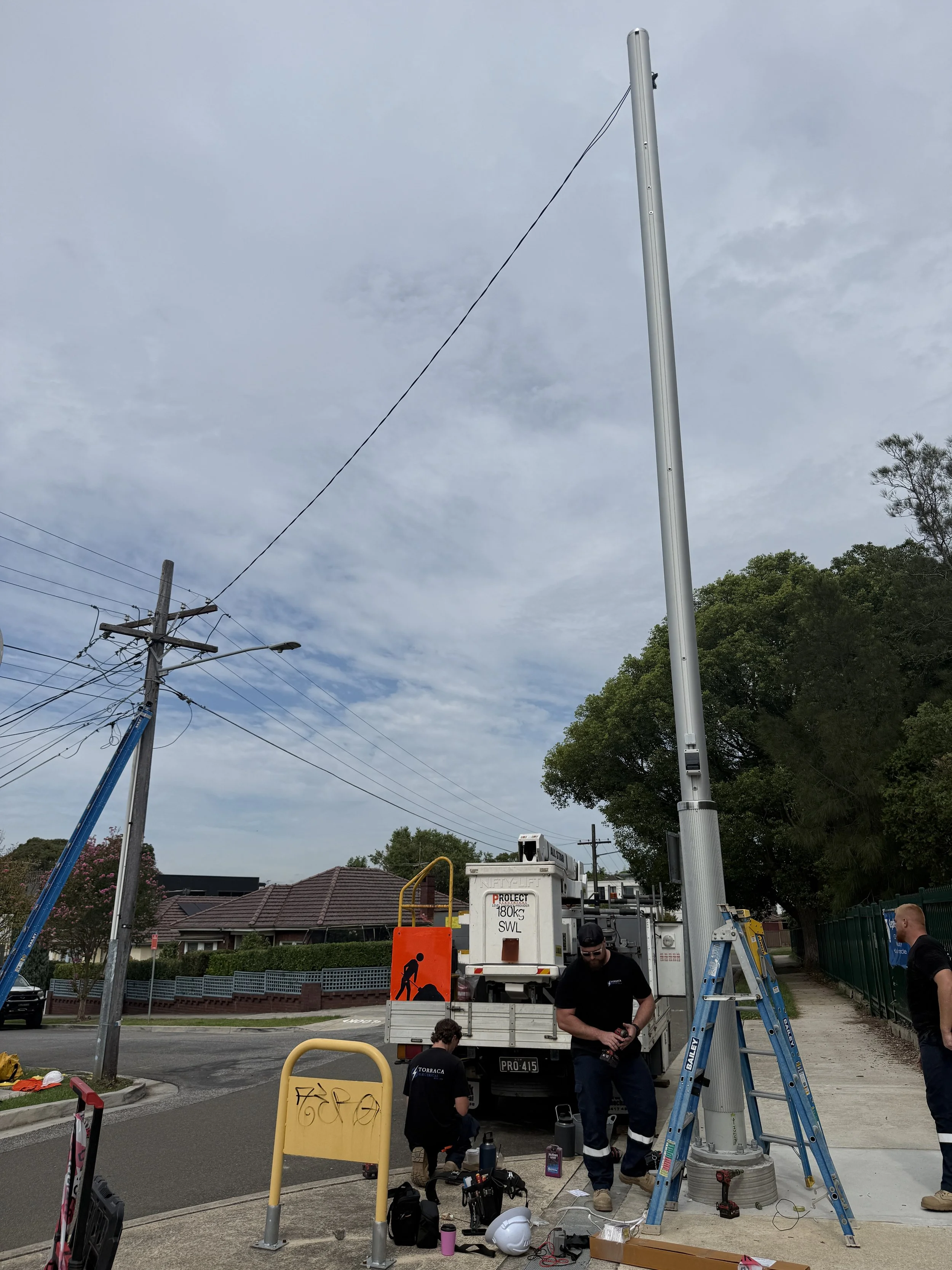 Workers installing a street light pole on a sidewalk with equipment and tools around them.