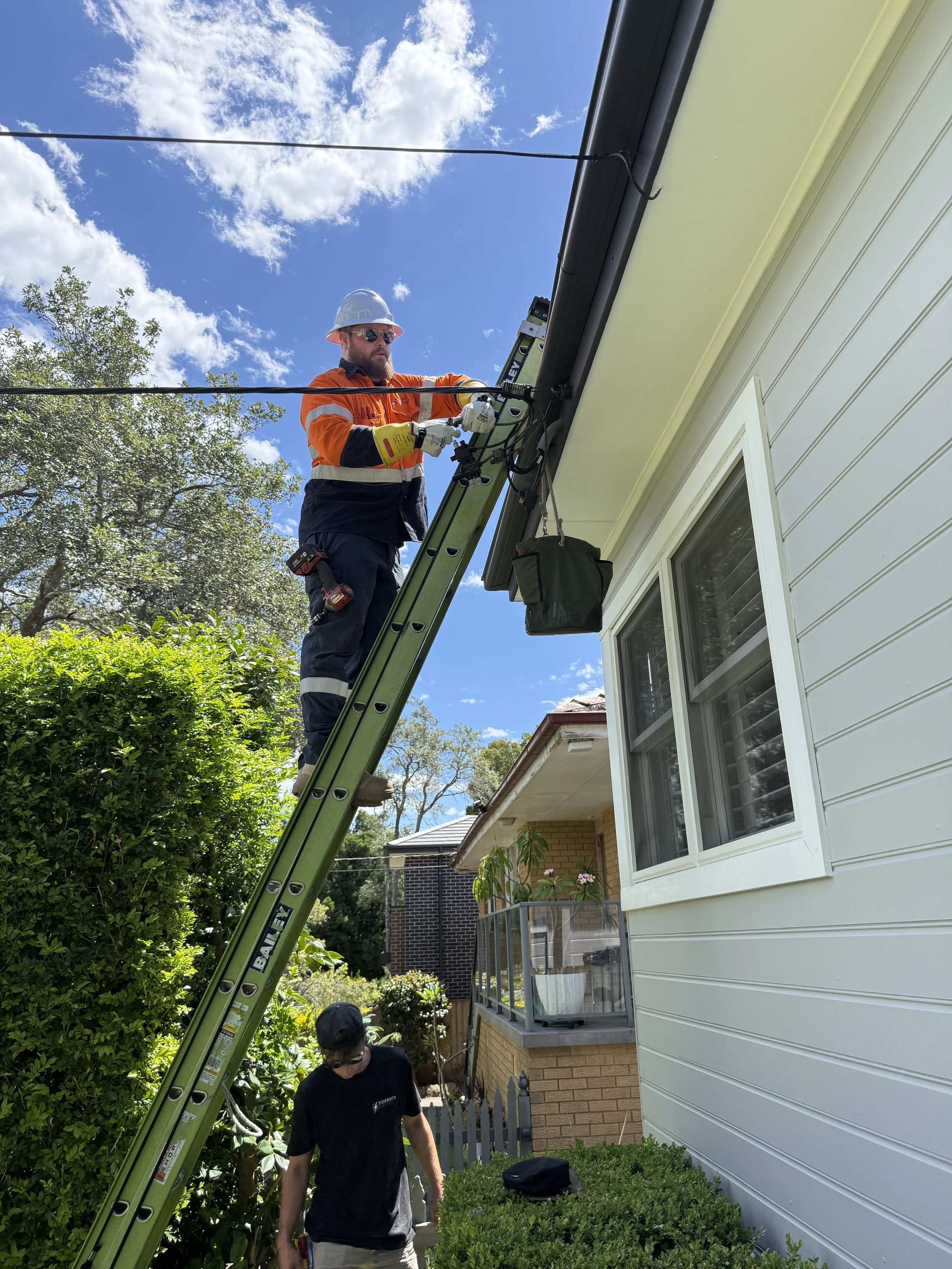 A worker in safety gear installing or repairing electrical lines on the side of a house, standing on a ladder with a second person below.