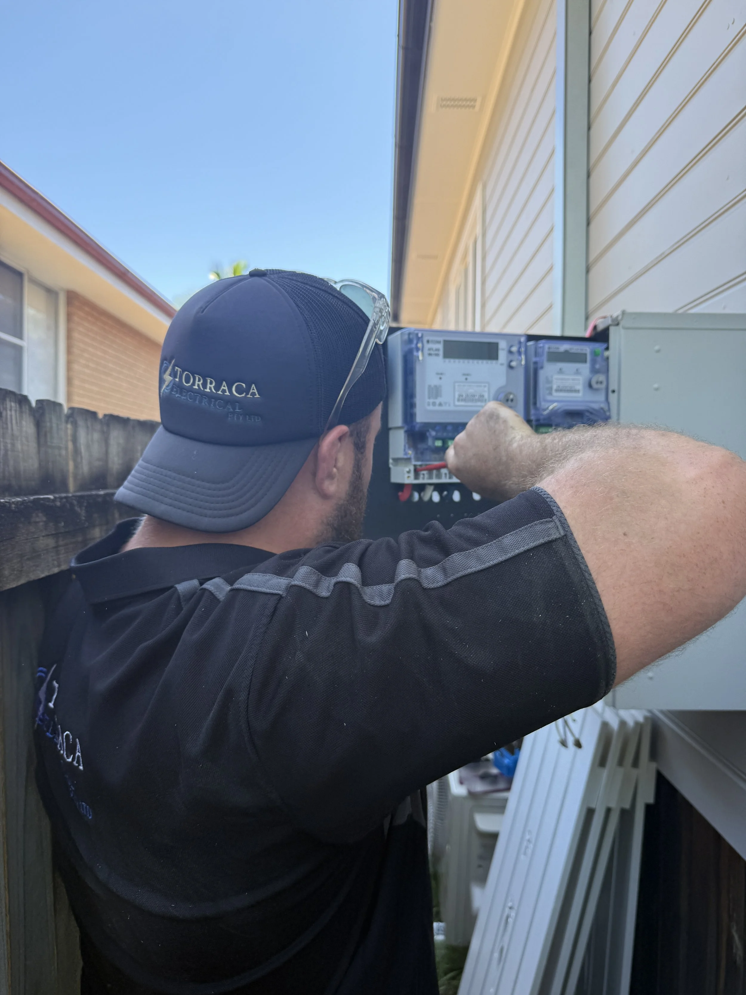 A technician working on an outdoor electrical meter box beside a house with a wooden fence and blue sky in the background.