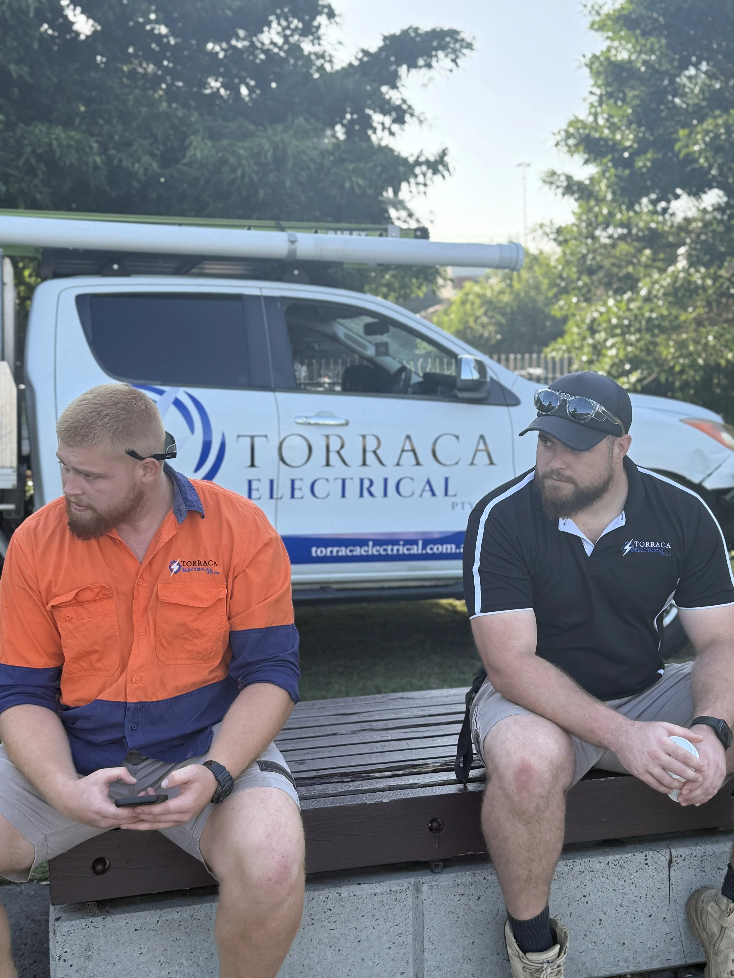Two men sitting on a bench outdoors, wearing uniforms with a Torra Electrical logo, with a Torra Electrical service truck in the background.