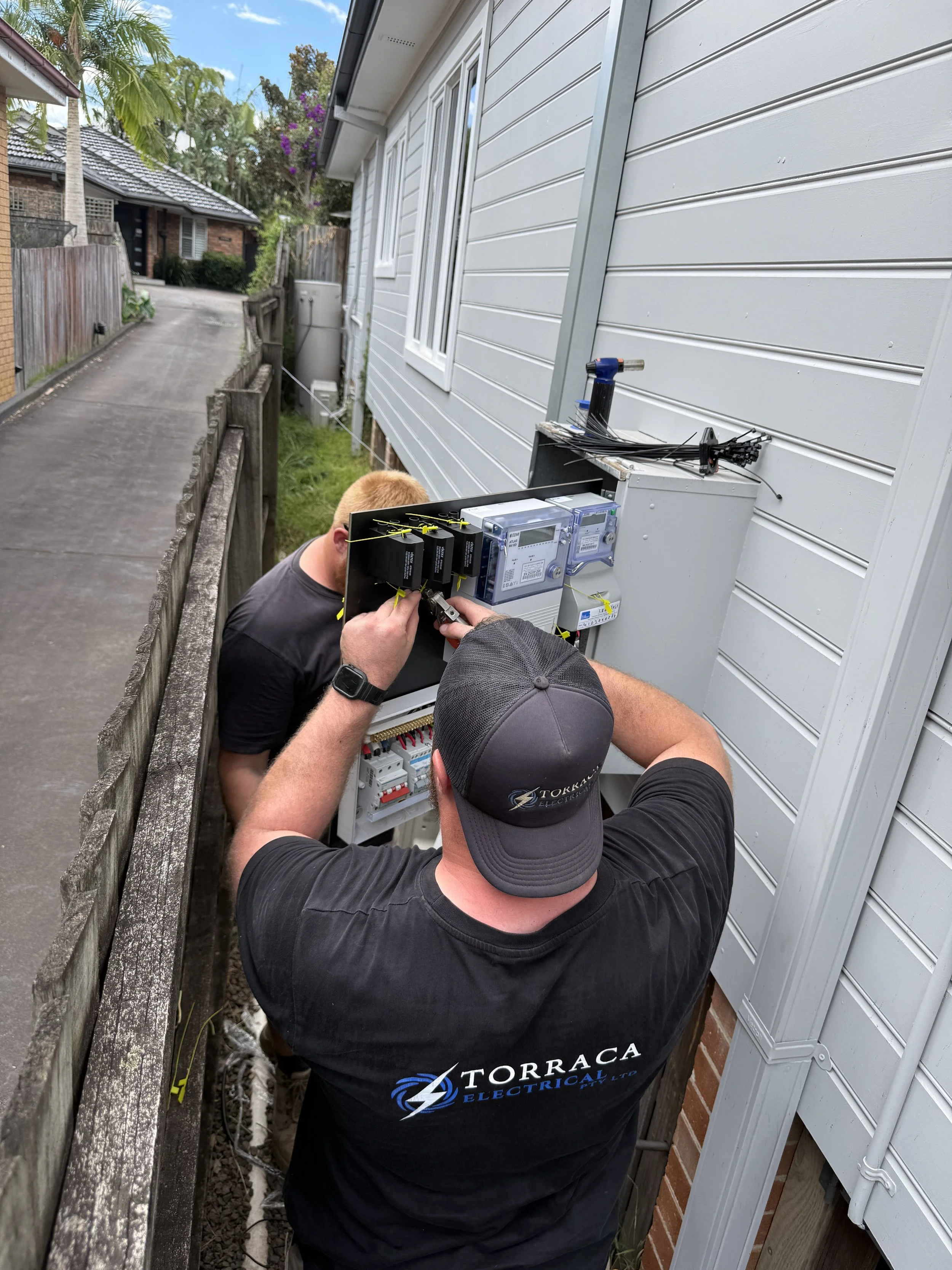 Two electricians working on an outdoor electrical panel beside a house, wearing black T-shirts with 'Torraca Electrical' logo, with a fence on the left and a suburban street in the background.