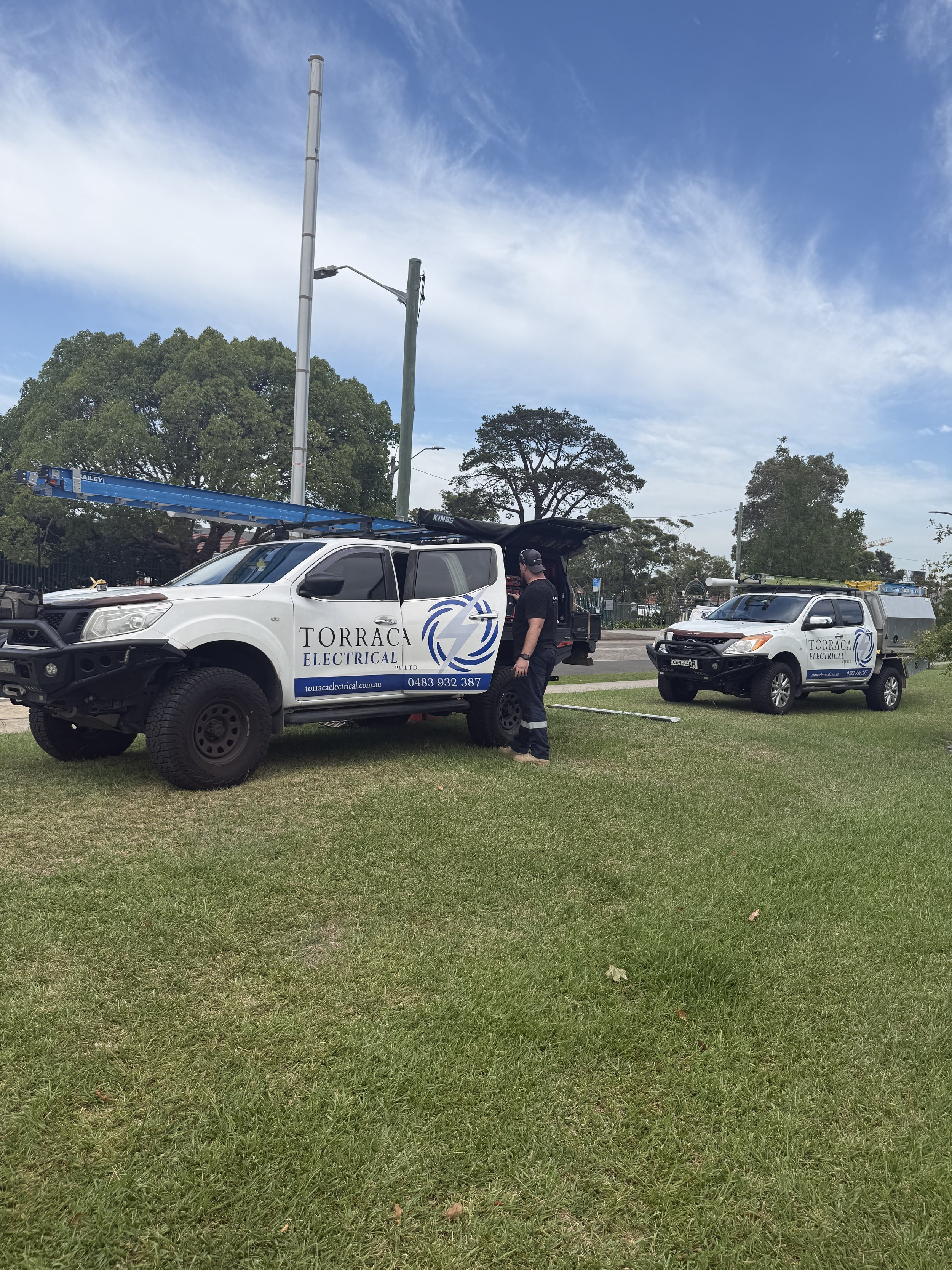 Two white utility trucks labeled 'Torraca Electrical' parked on a grassy area, with a technician working near the front of the trucks. Power lines and trees are visible in the background, under a partly cloudy sky.