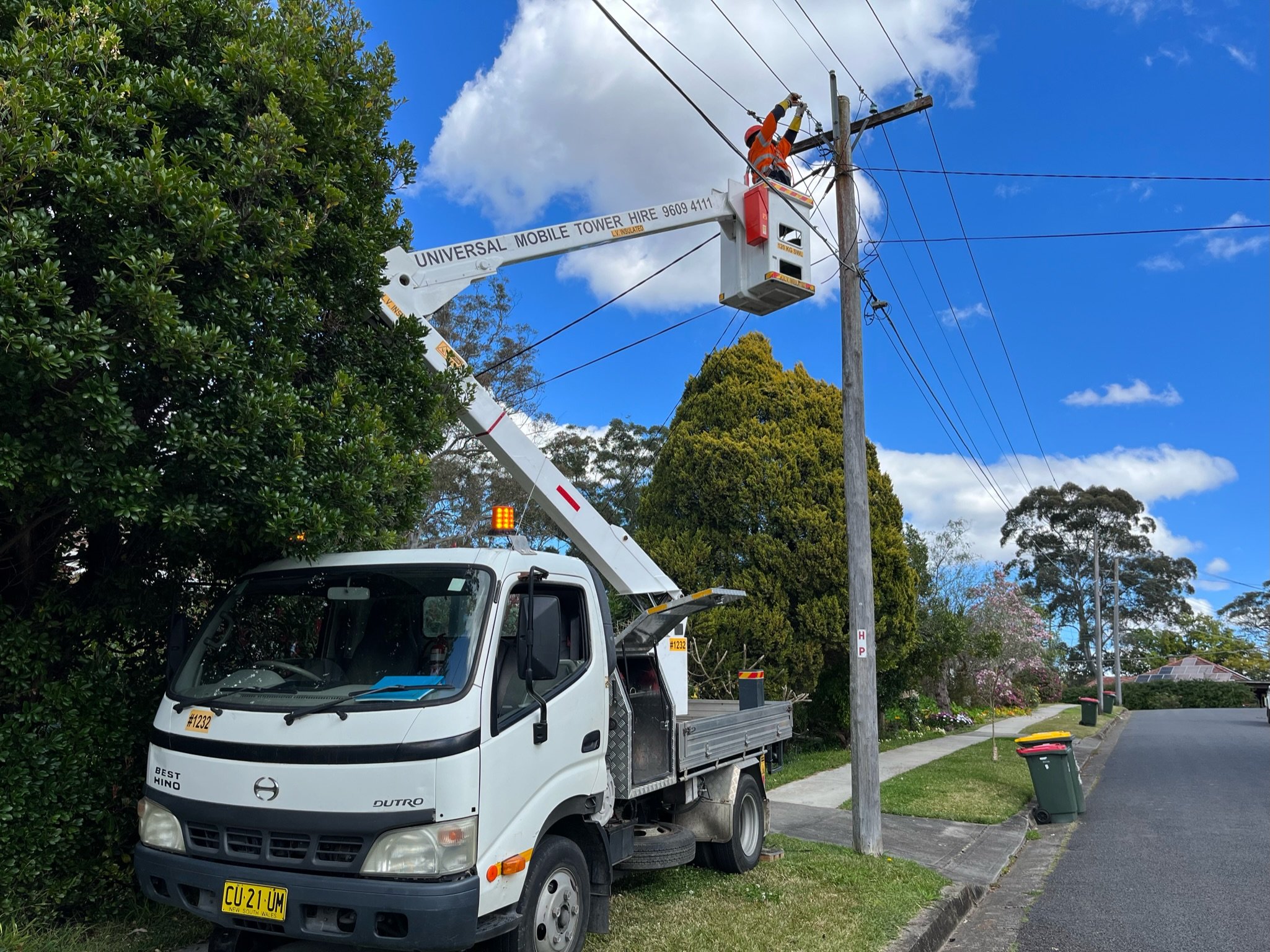 A utility worker in orange safety gear is on an elevated bucket lift trimming trees near an electrical pole on a suburban street. The lift is attached to a white truck parked on the grass next to the sidewalk. The sky is partly cloudy with some blue 