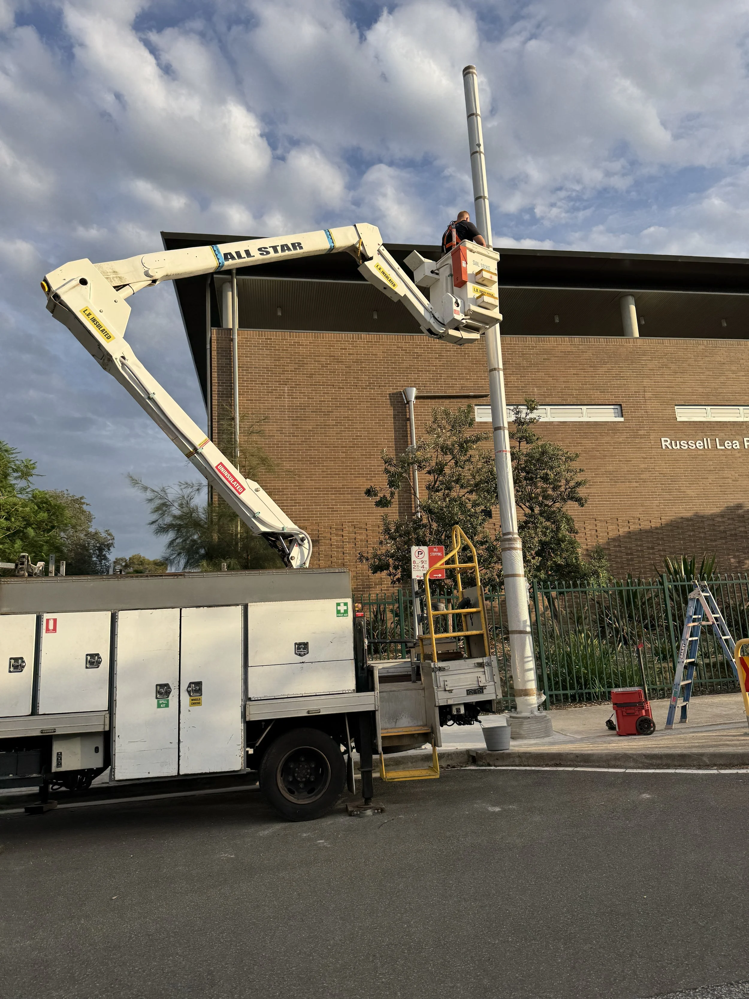 A worker in a bucket lift is repairing or maintaining a streetlight pole on a city sidewalk. The lift is attached to a truck parked on the street, with a building and cloudy sky in the background.