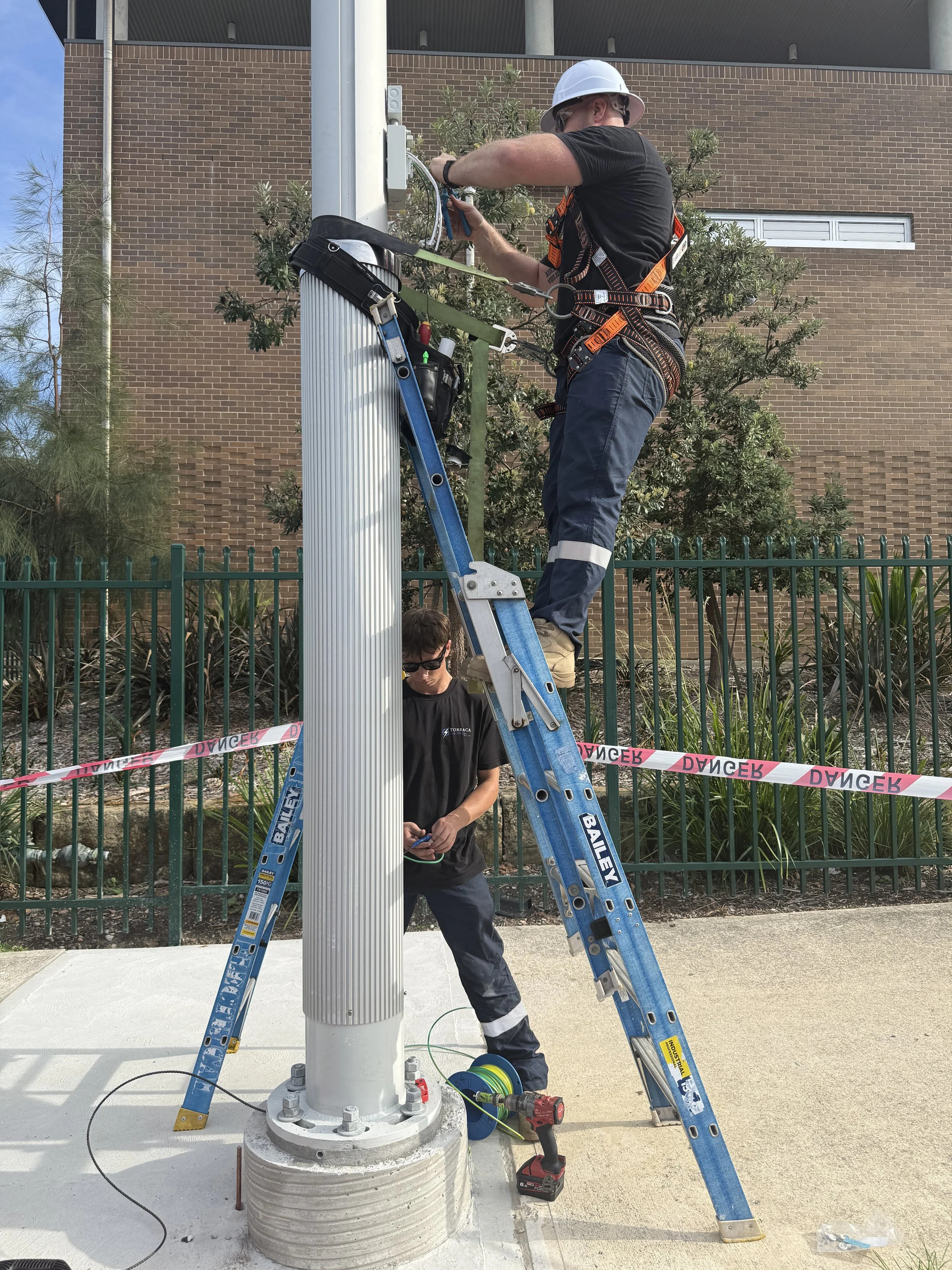 Two workers installing or maintaining a pole or electrical equipment outside. One worker is on a ladder working on the pole, while the other stands at the base, holding tools, with safety equipment and a drill nearby.