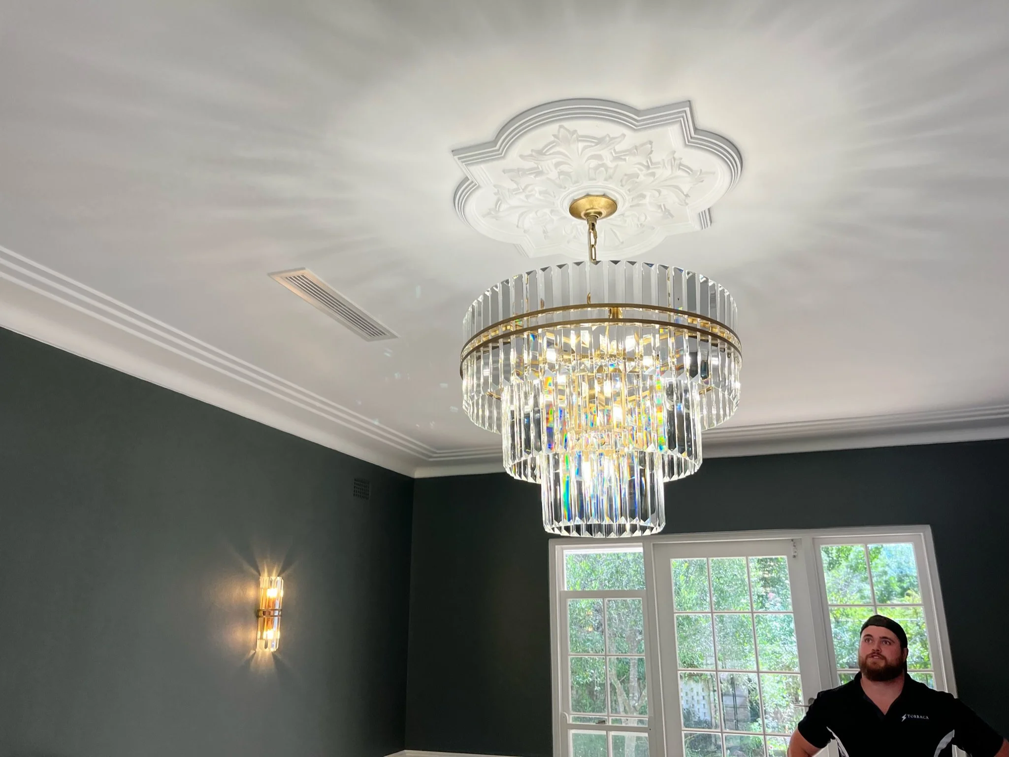 A bright, elegant chandelier with cascading crystal prisms hangs from a decorative ceiling medallion in a room with dark green walls and large window showing trees outside. A man stands near the window.