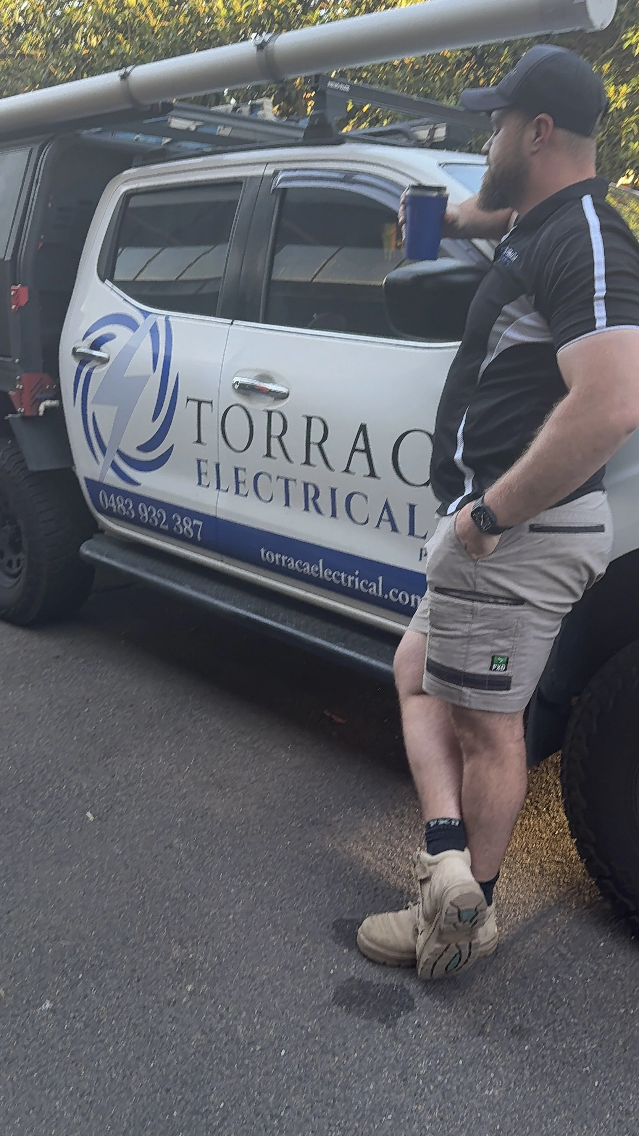 A man in casual work attire, including beige shorts, a black cap, and beige boots, standing beside a pickup truck with sign for Torraco Electrical. The man is holding a cup in his right hand and has his left hand in his pocket.