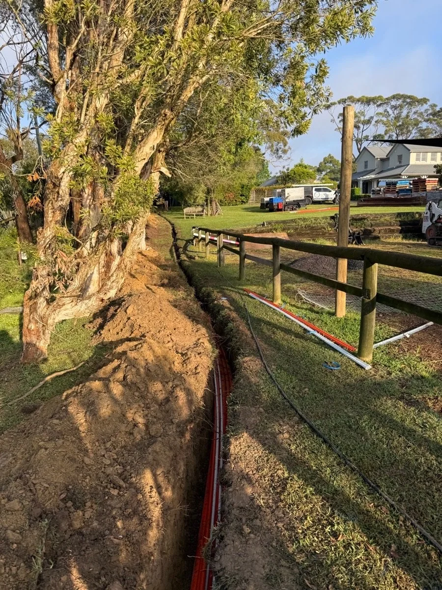 A trench with multiple orange cables installed along the side of a grass field, with trees and residential buildings in the background.