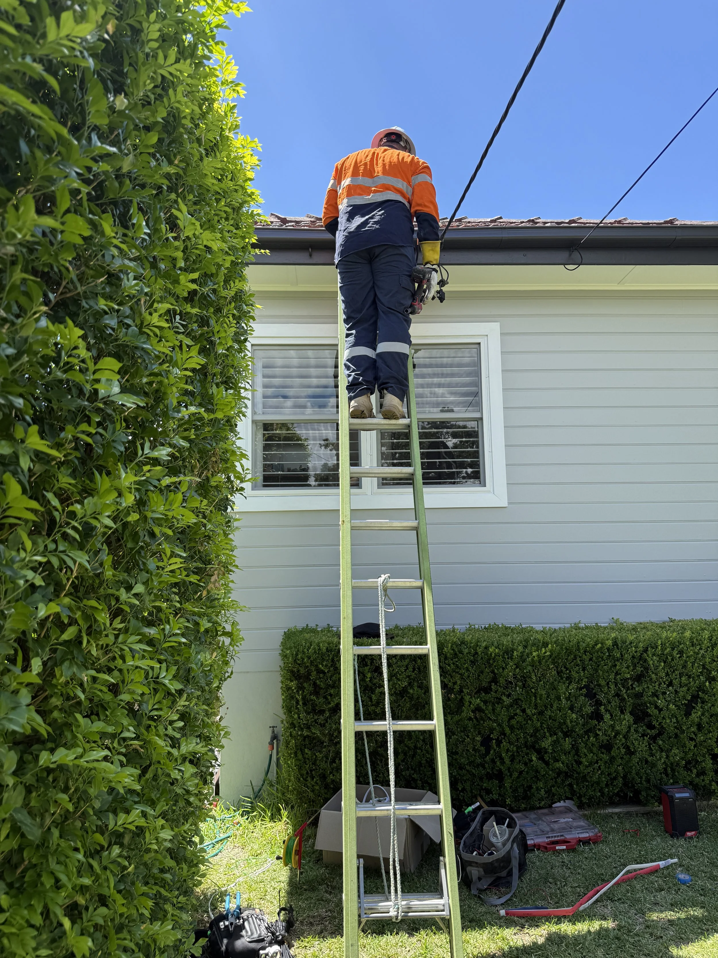 A worker in safety gear, including a helmet and orange safety vest, standing on a tall ladder and working on the roof of a house.